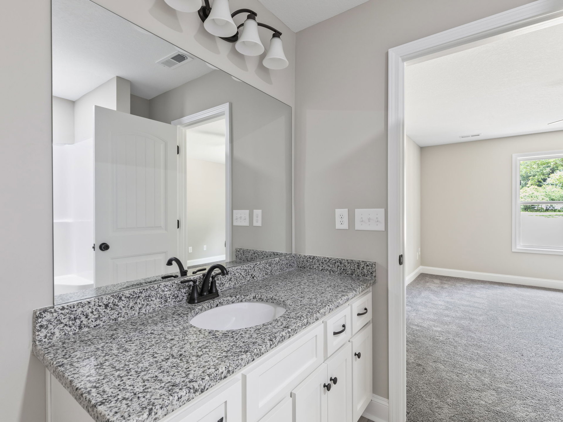 Bathroom featuring a marble countertop with a white sink and black faucet, large mirror above, three-light fixture with white lamps, white light switch, window revealing trees