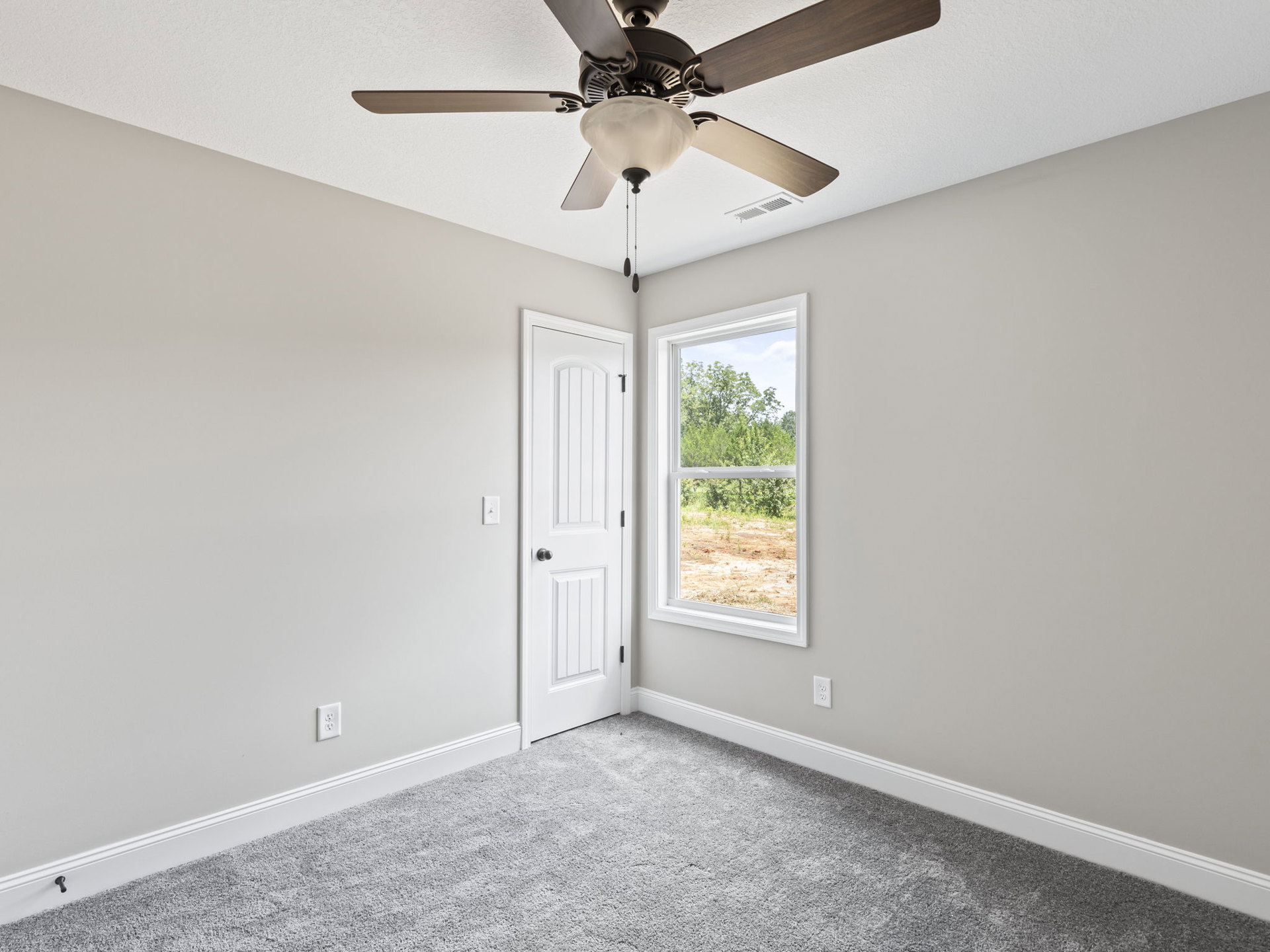 Ceiling fan with light fixture mounted above carpeted floor, white walls, open white door, and window showing green trees outside