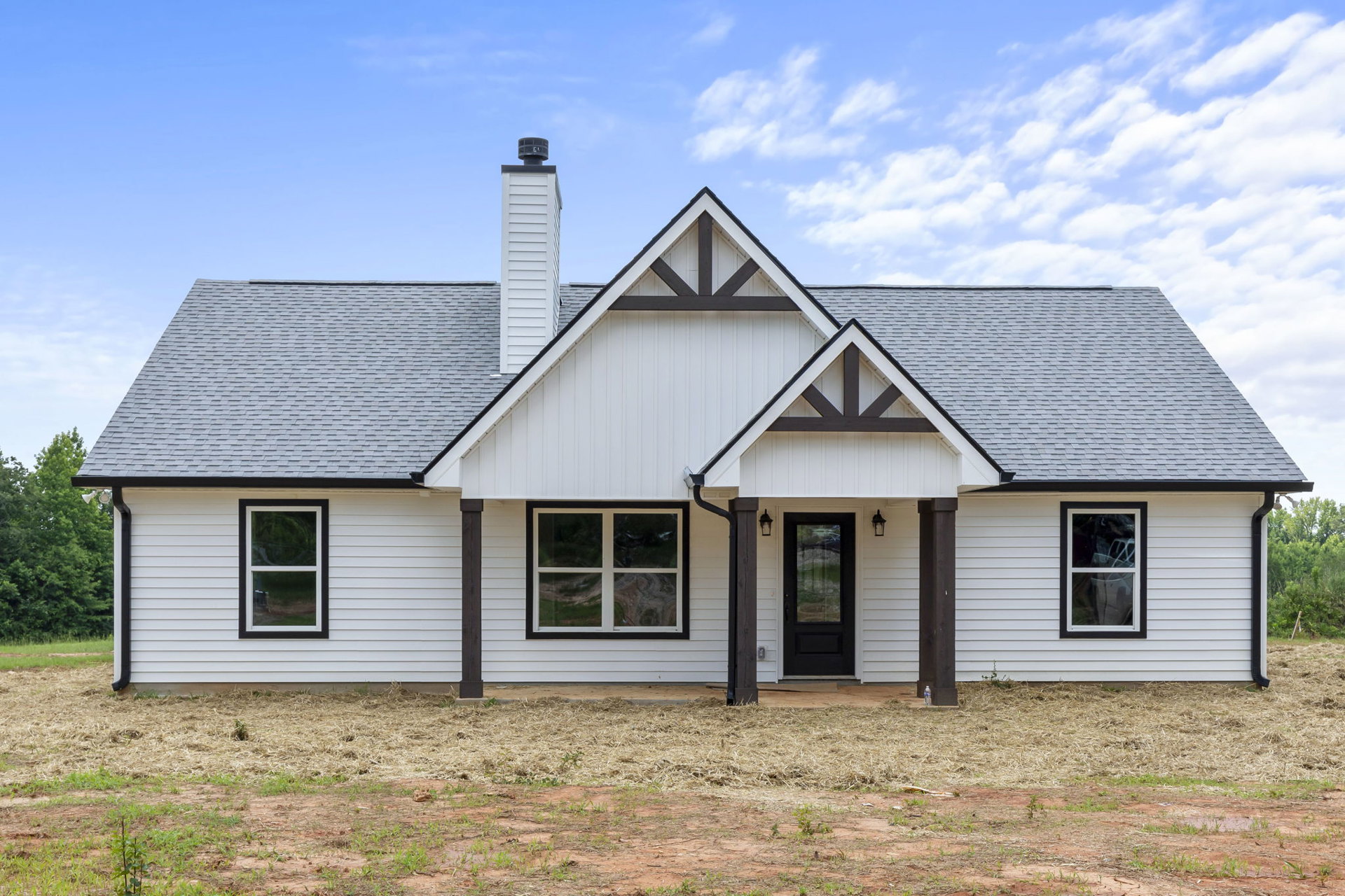 White siding house with black front door, white-framed window reflecting a car, hay pile on ground, cloudy sky above.