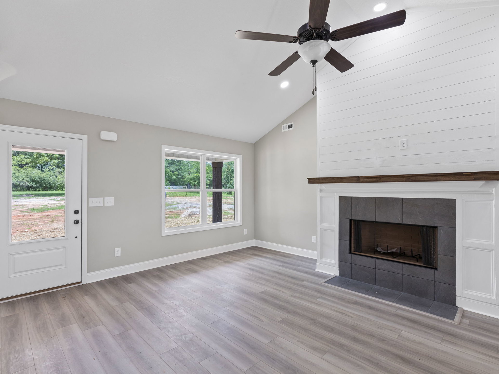 Living room with hardwood floors, stone fireplace featuring a metal rack, ceiling fan with light fixture, large window overlooking fenced yard, and white door opening to outdoor