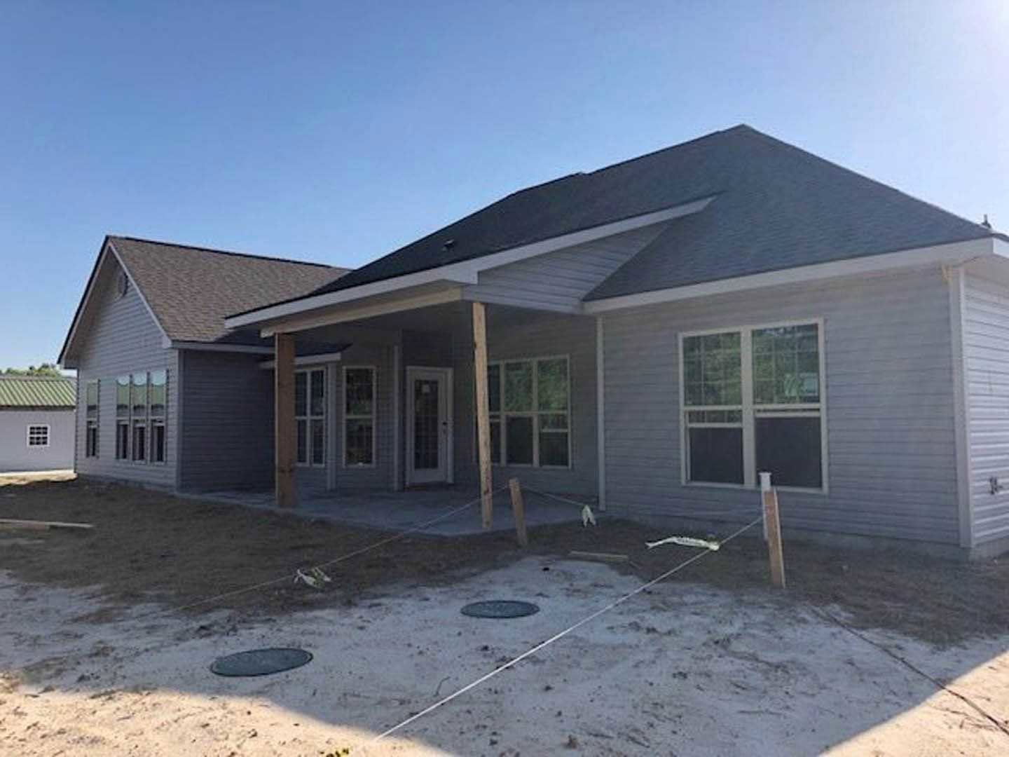 Two-story house under construction with white-framed windows, grid window, covered porch, wood siding, fenced yard, and manhole cover in dirt foreground