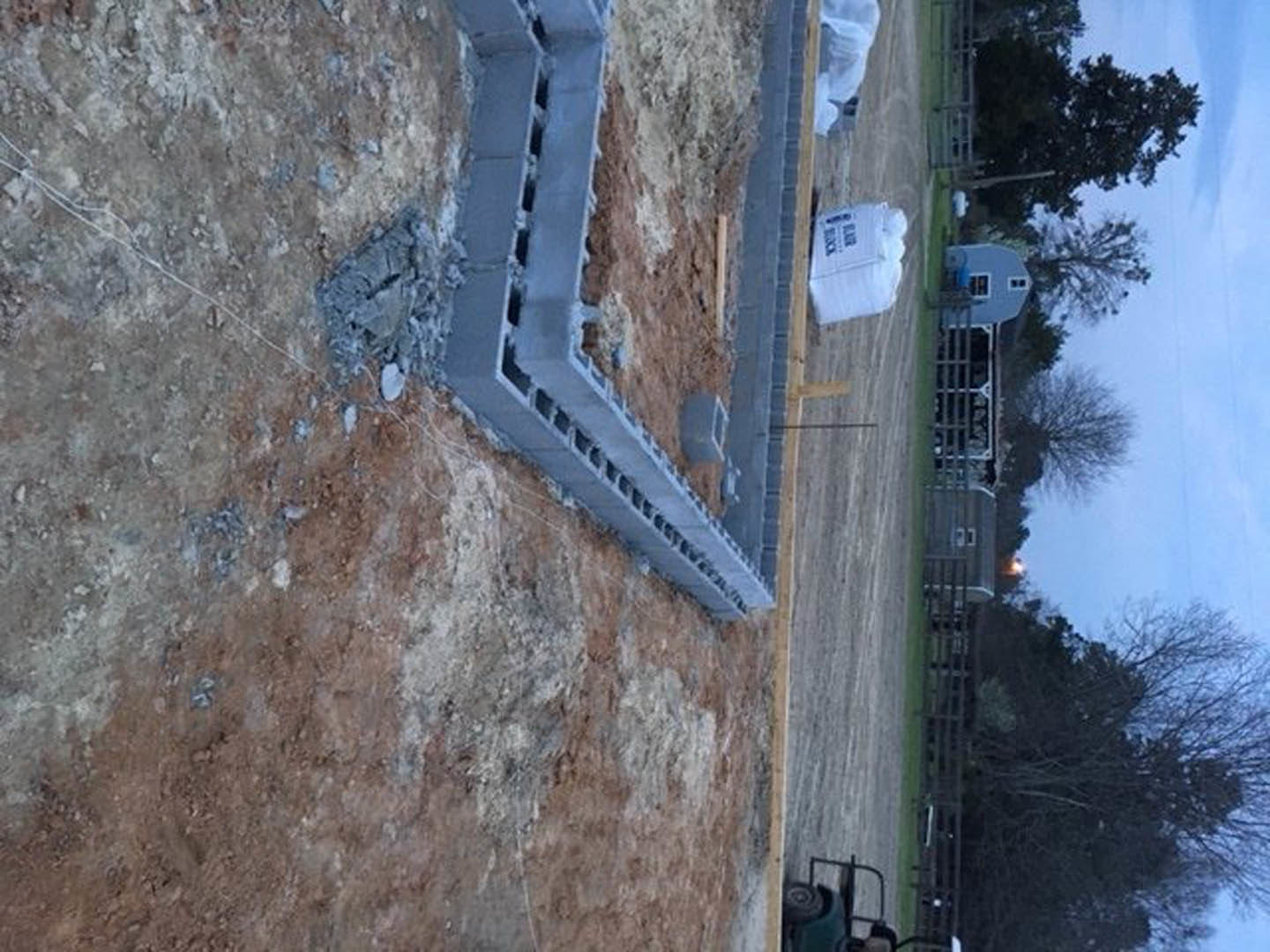 Partially built home foundation surrounded by dirt and construction materials, fenced perimeter, open grassy field and cloudy sky in background.