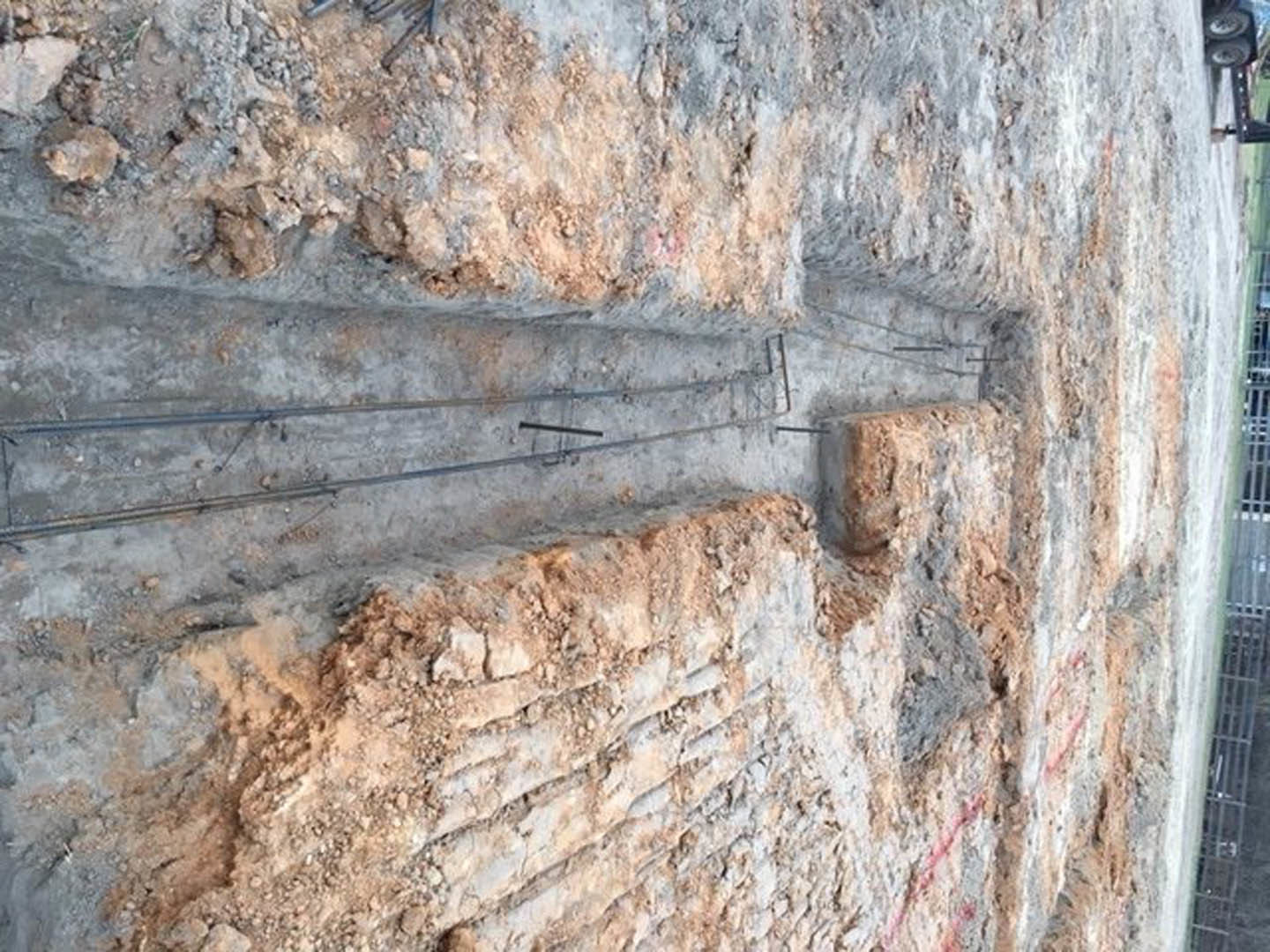 Limestone exterior wall under construction with exposed metal rods, surrounded by dirt and large rocks