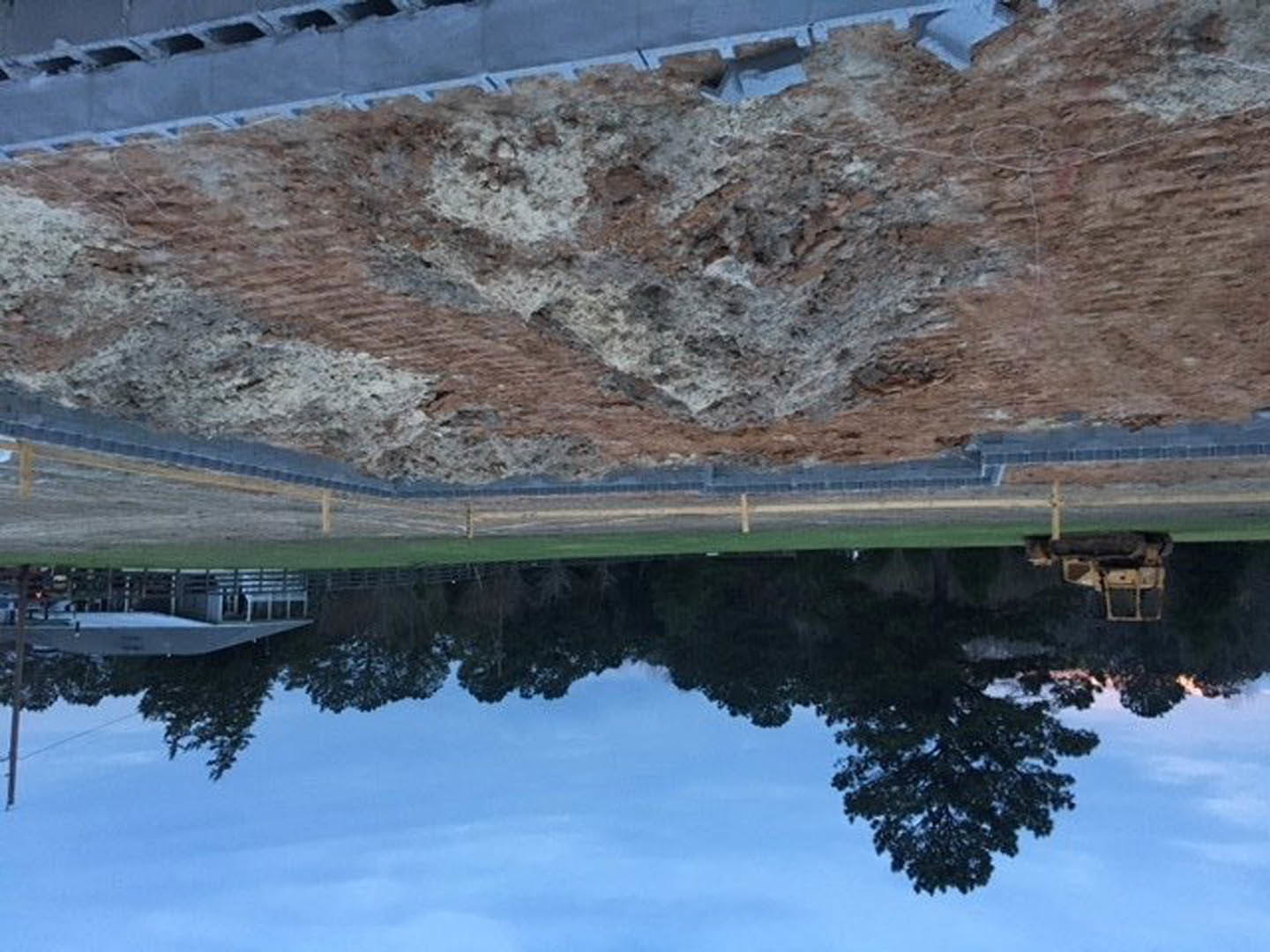Framed construction site beside a lake, unfinished wooden structure, exposed beams, water reflecting sky, trees lining shoreline in background