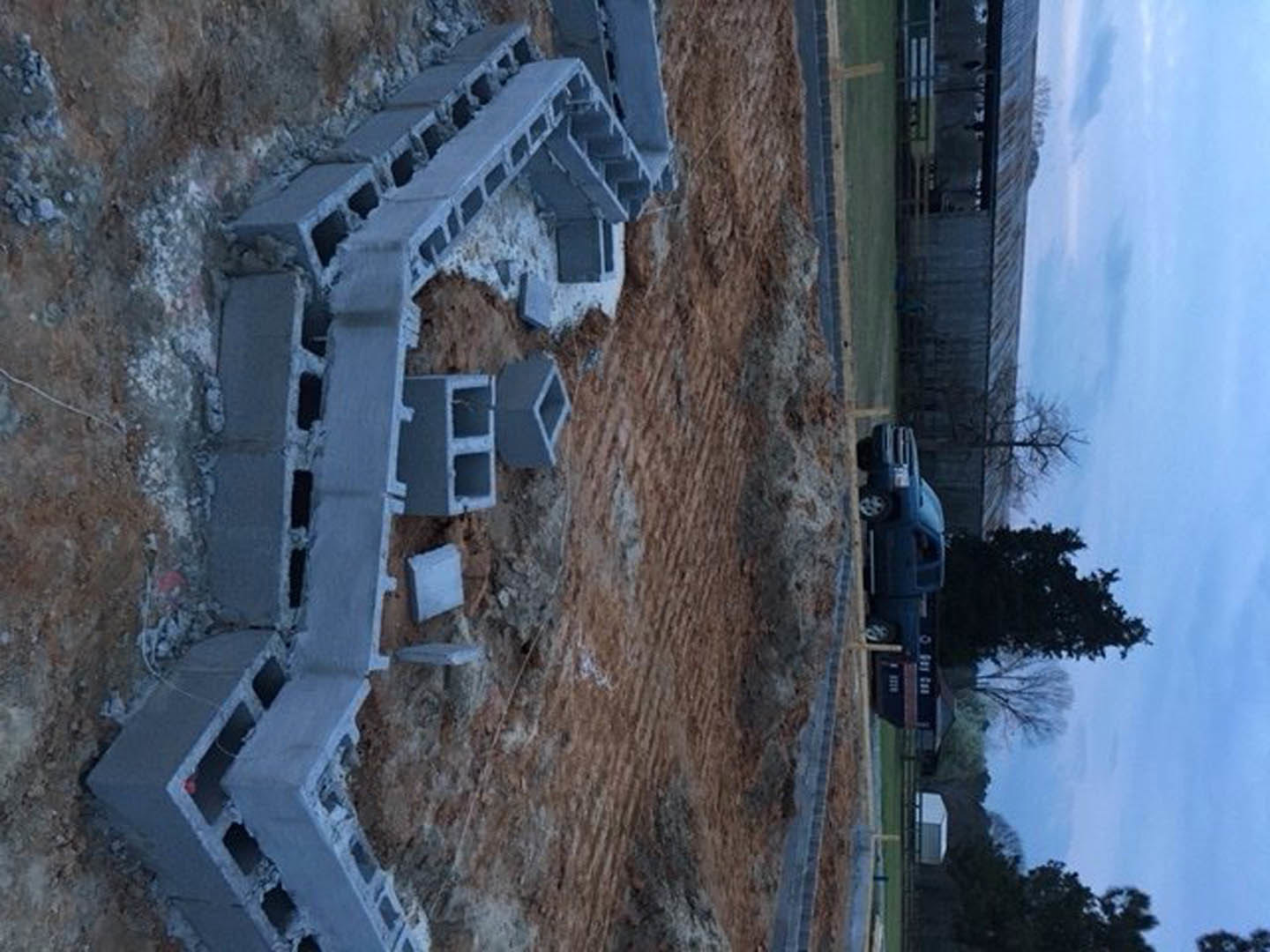 Partially built custom home with exposed concrete blocks, construction truck parked in front, scattered materials on dirt lot, mature tree beside structure, cloudy sky overhead