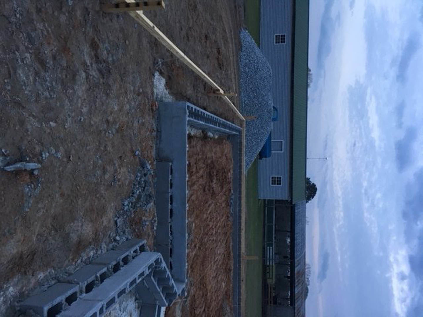 Stone wall and concrete stairs leading to unfinished custom home exterior under blue sky with scattered clouds, dirt construction area in foreground.
