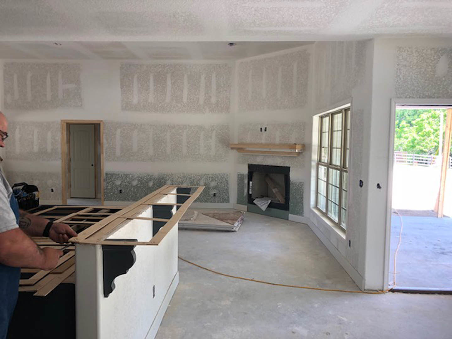 Man assembling wood trim near white door with black handle, plaster walls, light wood flooring, and kitchen cabinetry visible in background