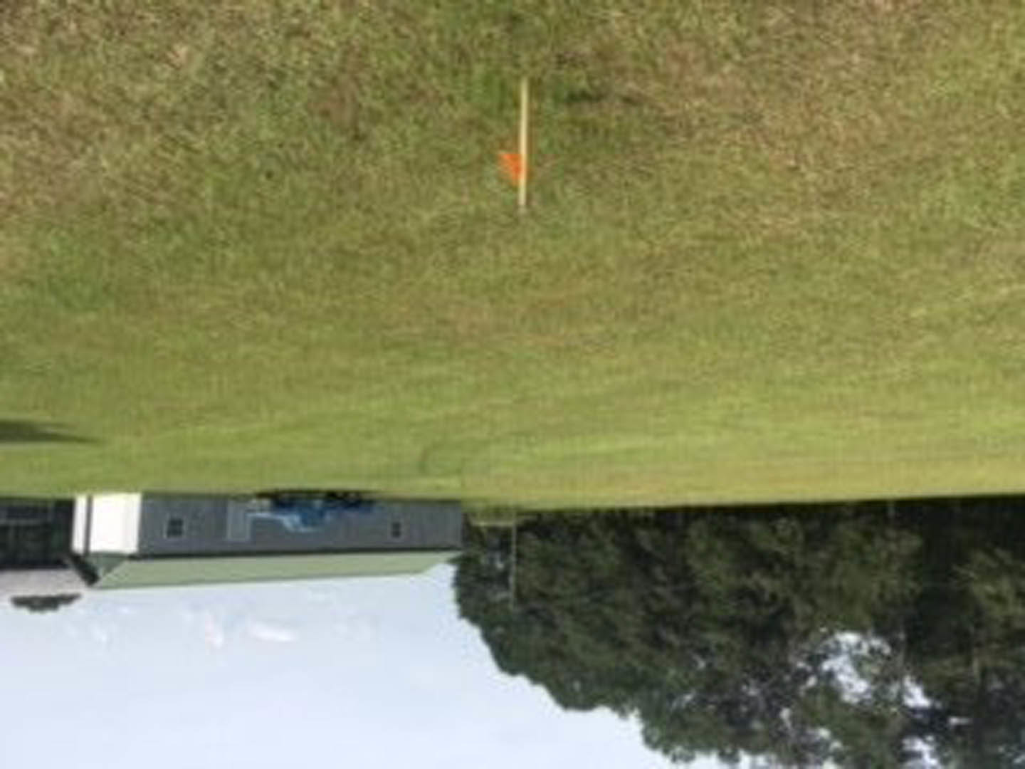 Modern home with light siding and large windows set beside a leafy tree, American flag on a pole in manicured grass, expansive field and lake in background