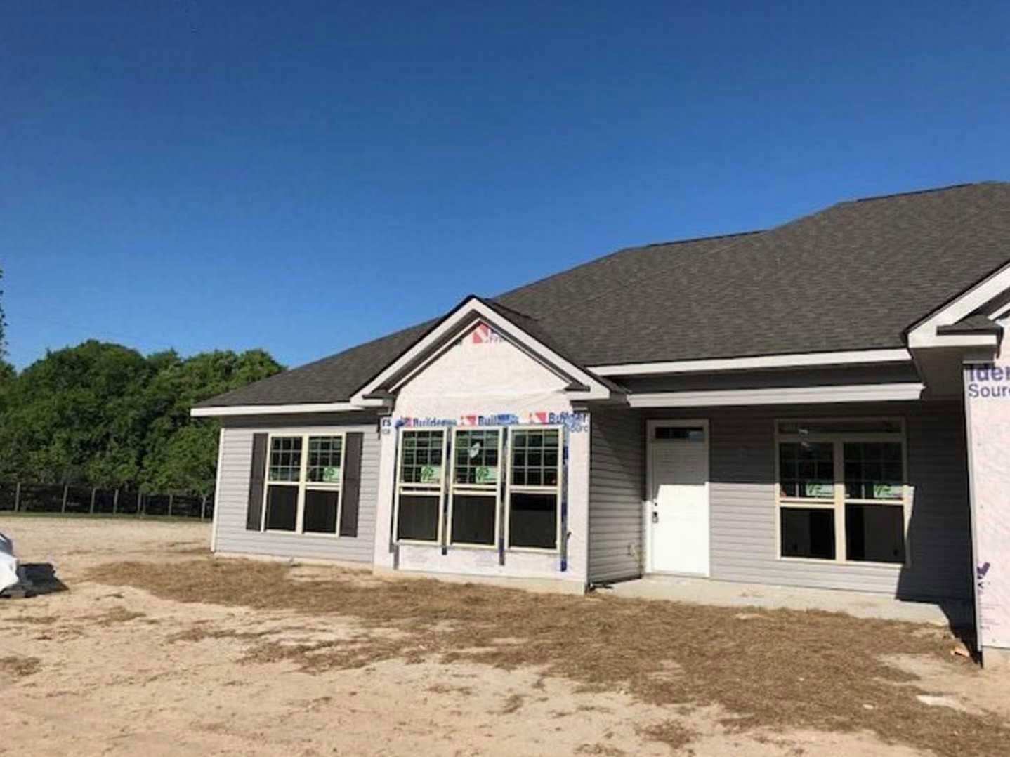 Two-story house with white door and windows, light-colored siding, dirt patch in front, grassy yard, and trees in the background