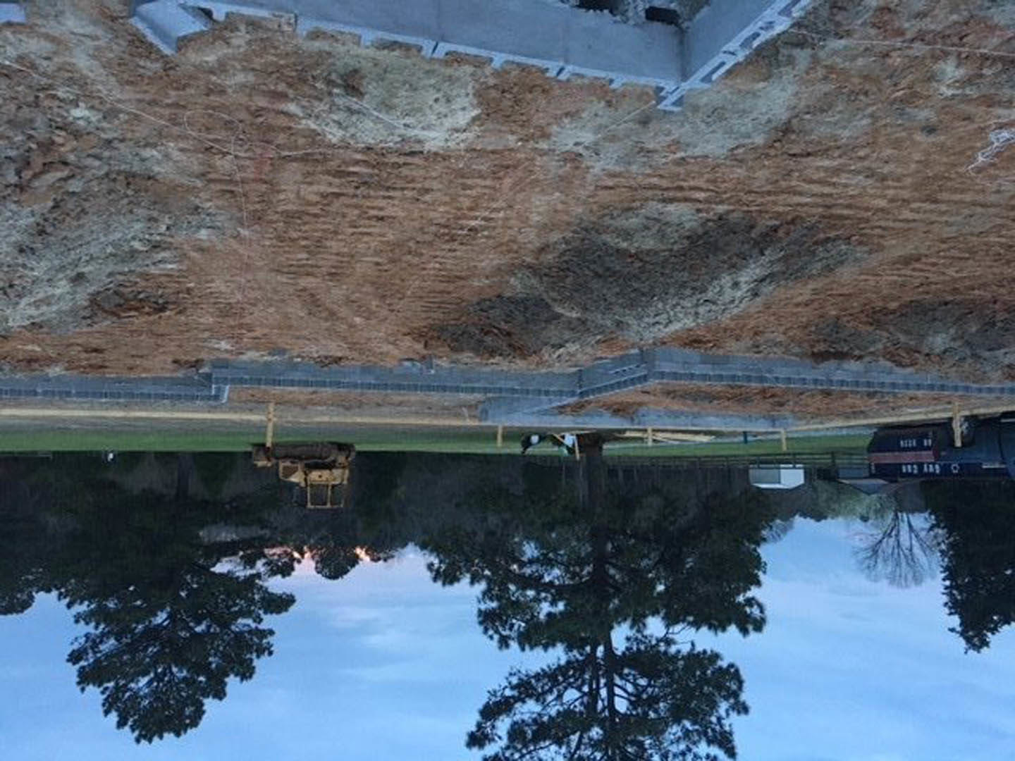 Large dirt hill with yellow tractor, sparse vegetation, tree reflected in nearby water, concrete wall with metal grate in background