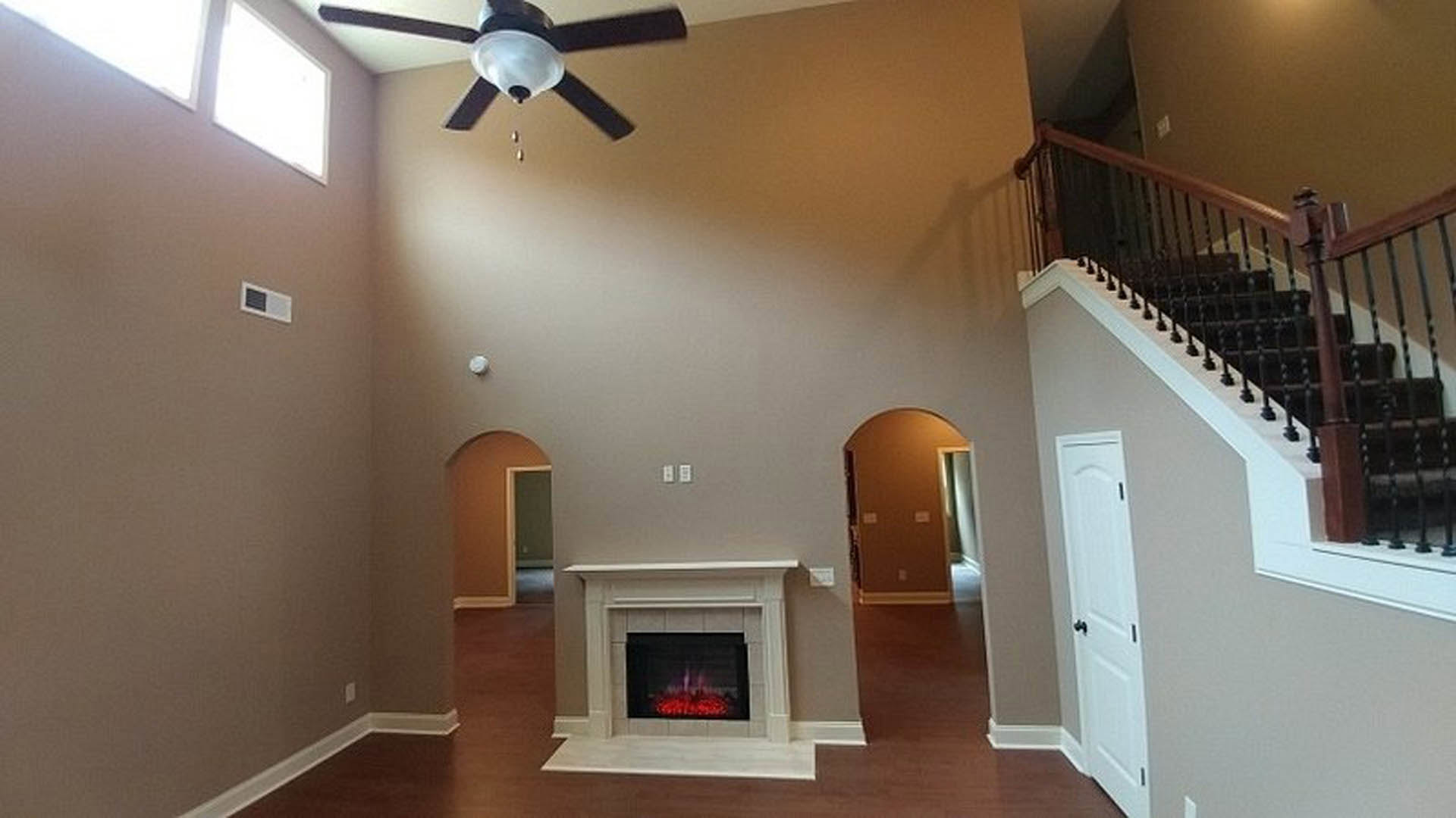 Stone fireplace with burning fire, white plaster walls, wood floor, ceiling fan, paneled door, staircase featuring built-in dog house under steps