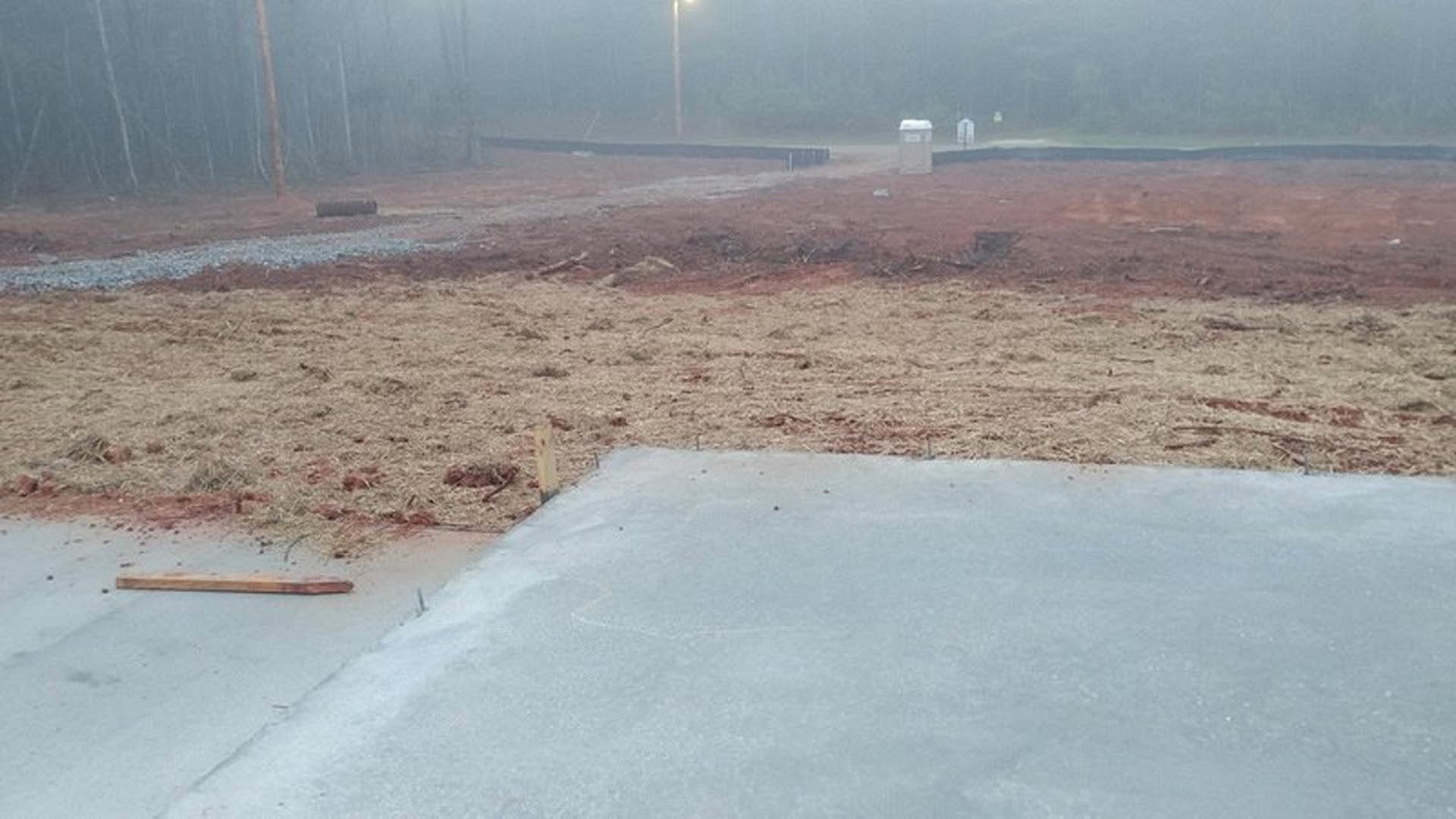Concrete slab foundation surrounded by dirt, wooden post visible, foggy outdoor setting with bare trees in background