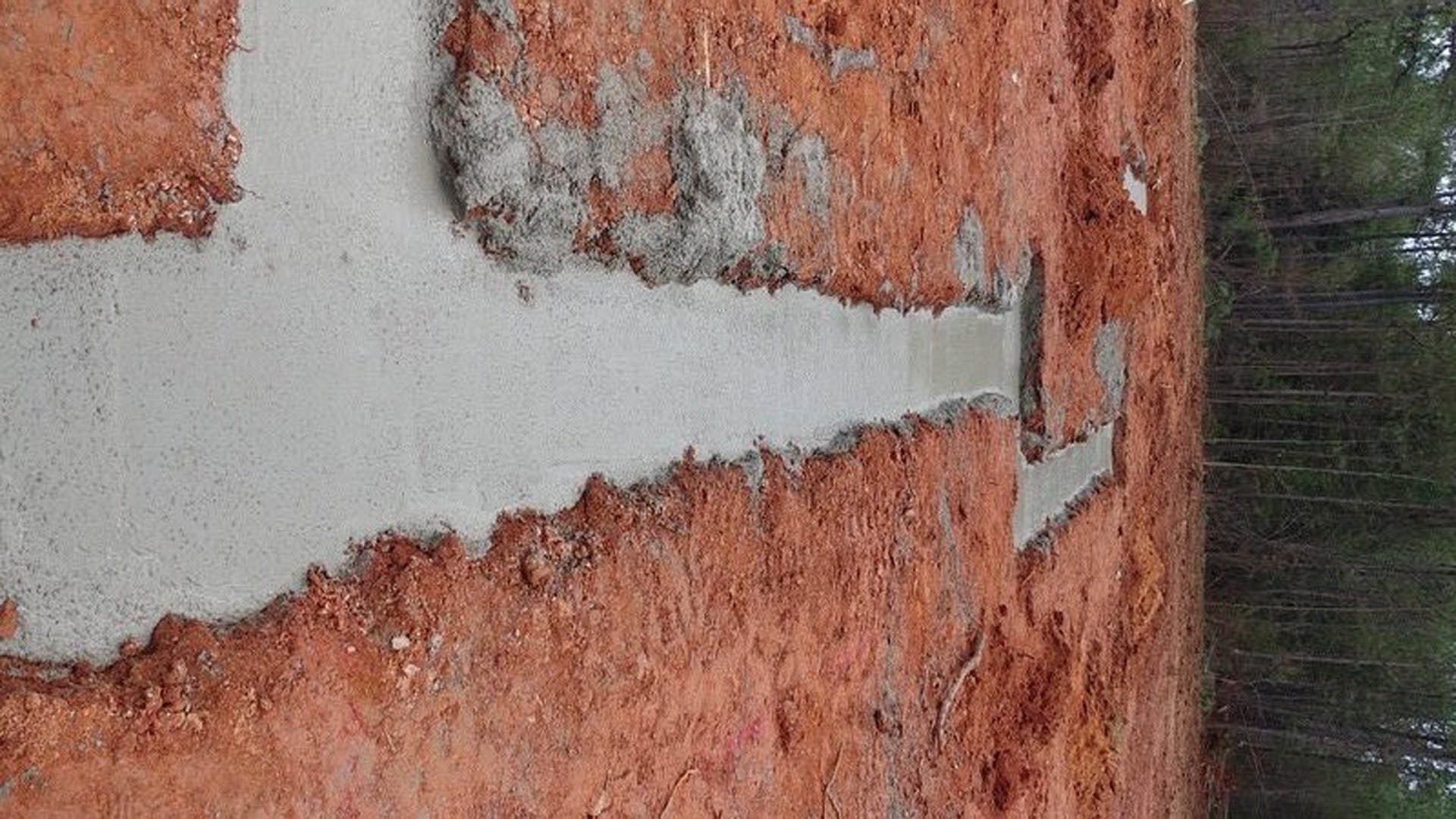 Freshly poured concrete slab foundation surrounded by red dirt, bordered by trees in the background