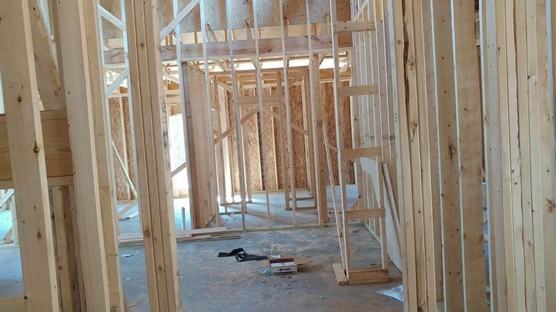 Exposed wood framing and beams in a partially constructed room, wooden ladder leaning against wall, cardboard boxes and black object on snow-covered floor, building insulation