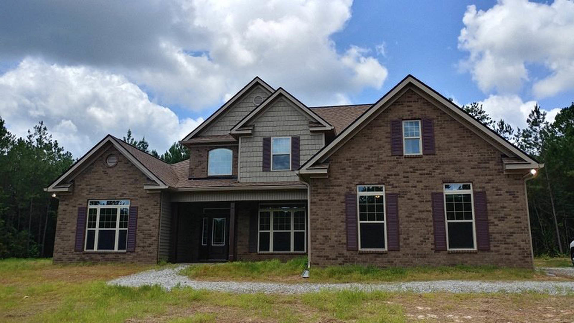 Two-story house with gray siding, white-framed windows, shingled roof, paved driveway, green lawn, and mature trees under a clear blue sky
