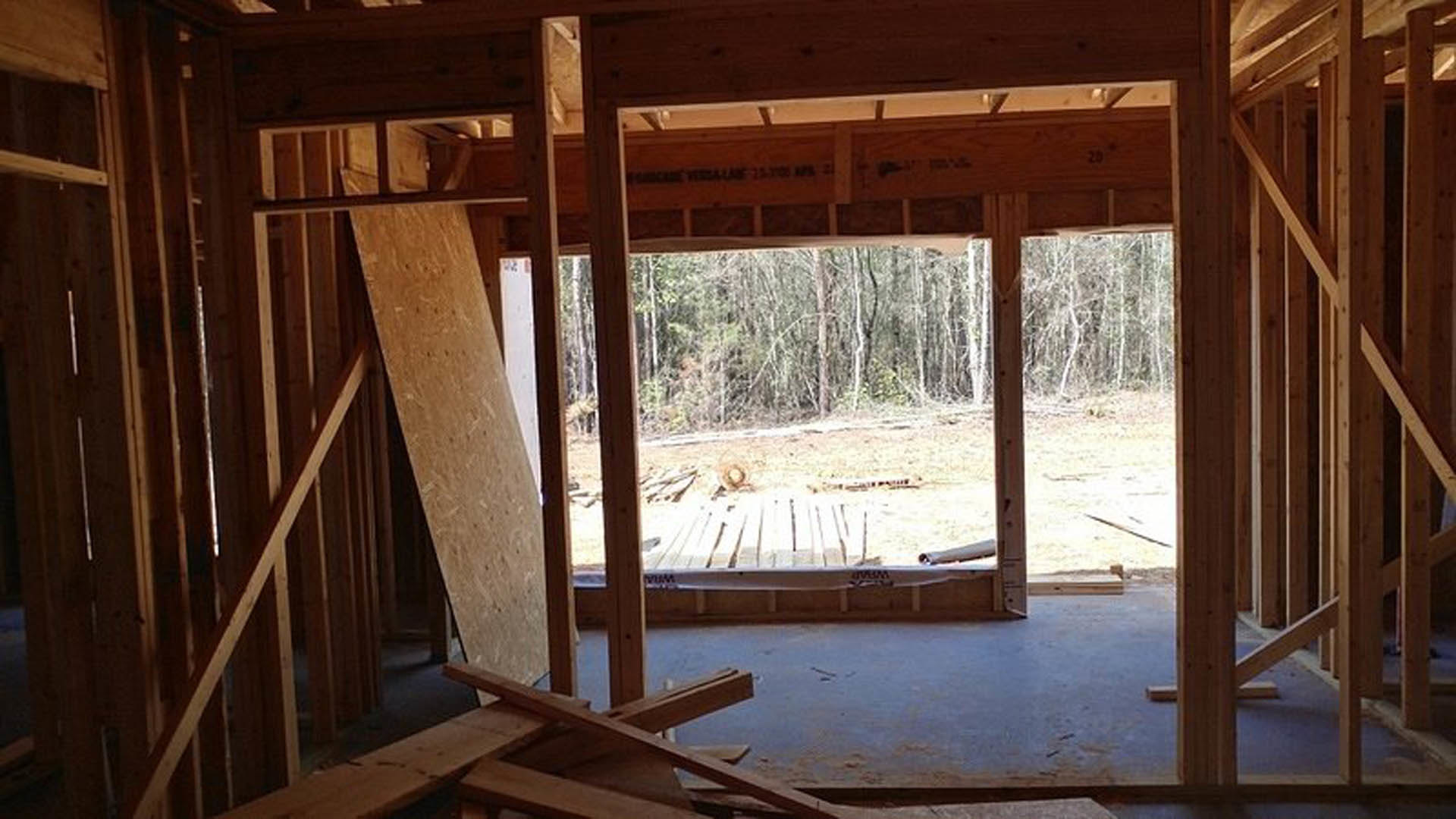 Sunlit room featuring wood plank flooring, exposed wooden beams, and a large window overlooking a forested landscape