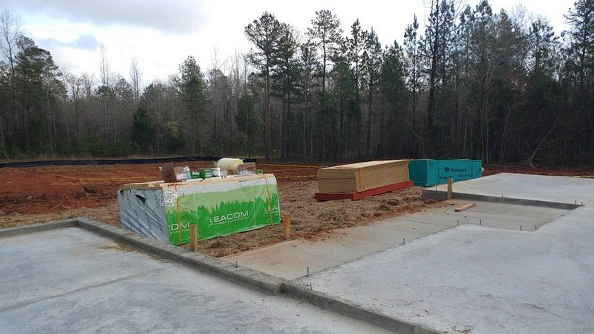 Framed construction site with stacked lumber, wooden pallets, green and white tarp with printed text, blue sign, and surrounding trees under cloudy sky