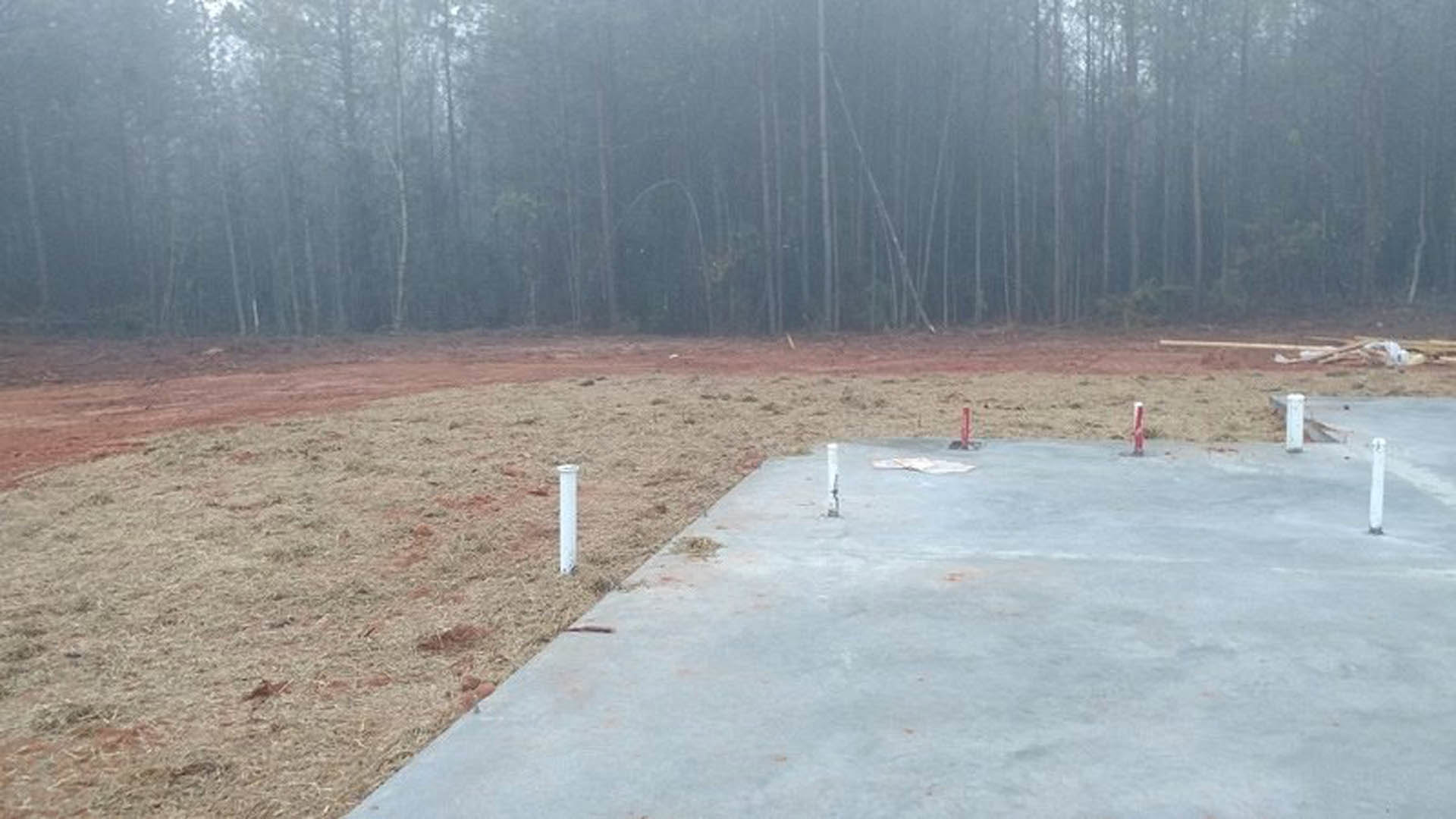 Concrete slab foundation with several upright poles set in front of a dirt field, surrounded by foggy trees in the background.