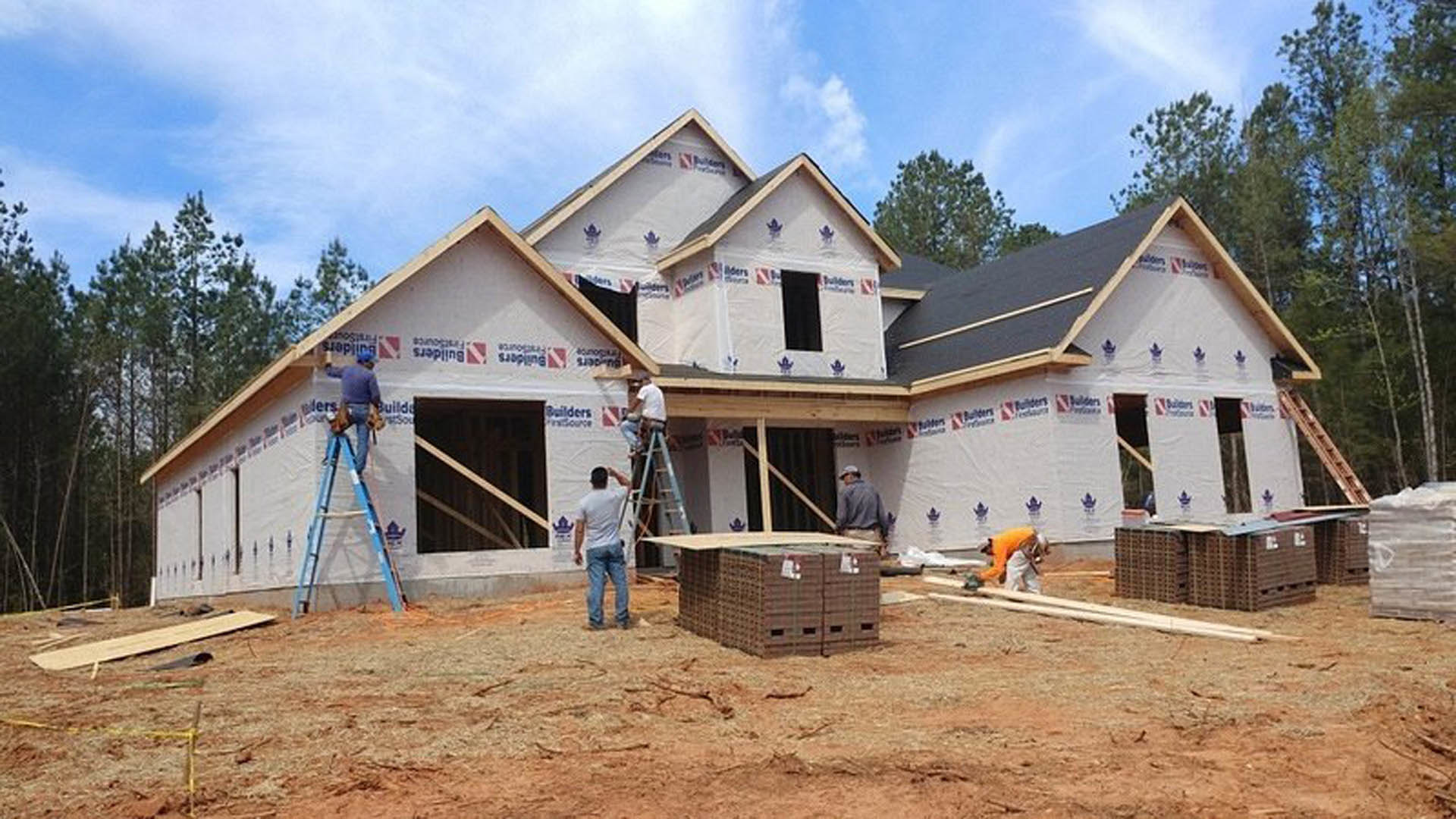 Men constructing a wood-framed house exterior, ladder propped against unfinished siding, scattered lumber on ground, cloudy sky overhead, trees in background