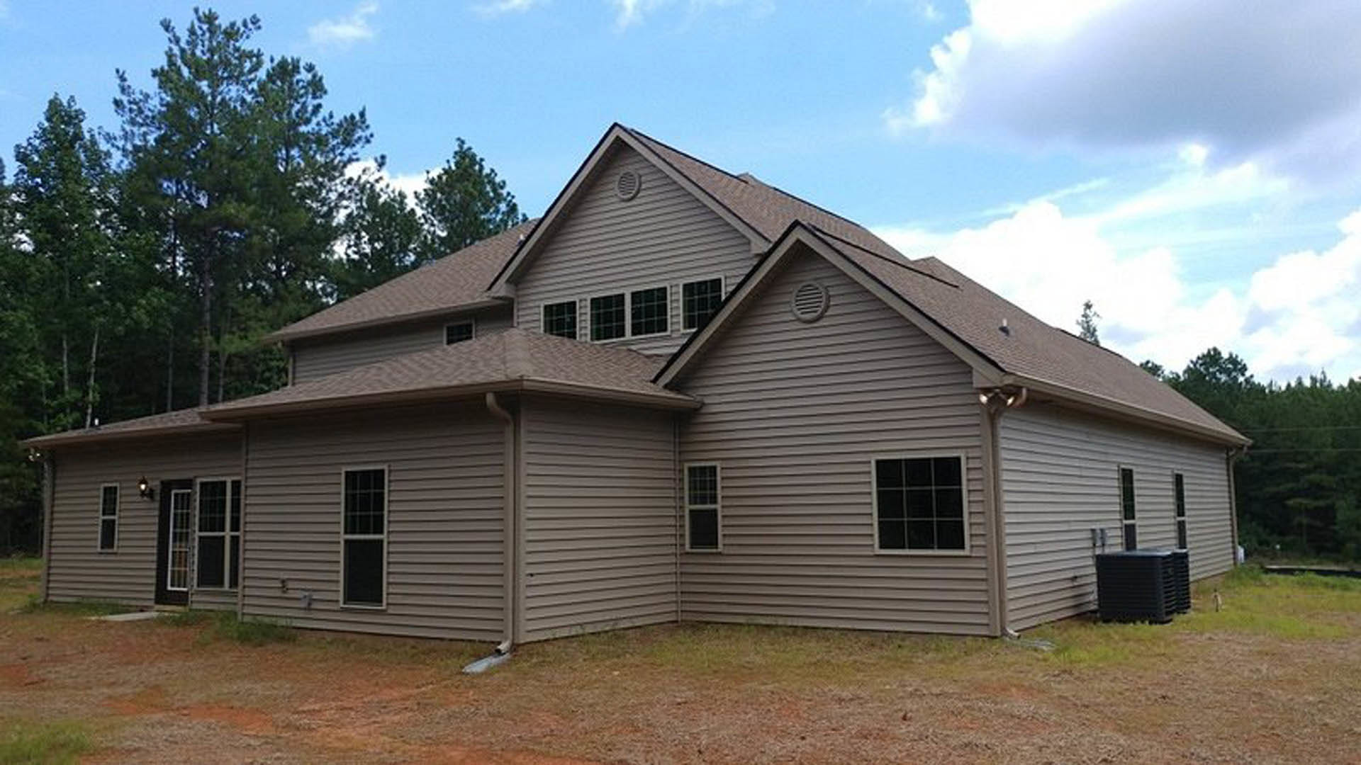 Modern home with expansive gabled roof, light-colored siding, multiple grid windows, paved driveway, and manicured lawn surrounded by trees under a cloudy sky