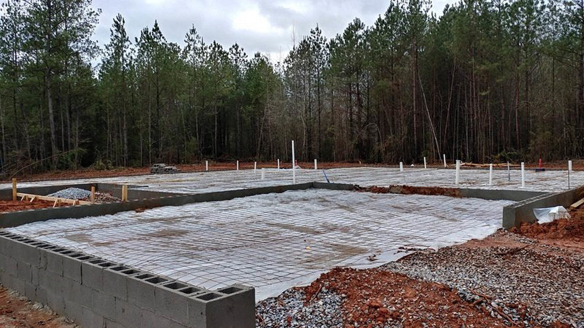 Concrete foundation slab bordered by a partial brick wall, surrounded by mature trees and greenery under a cloudy sky