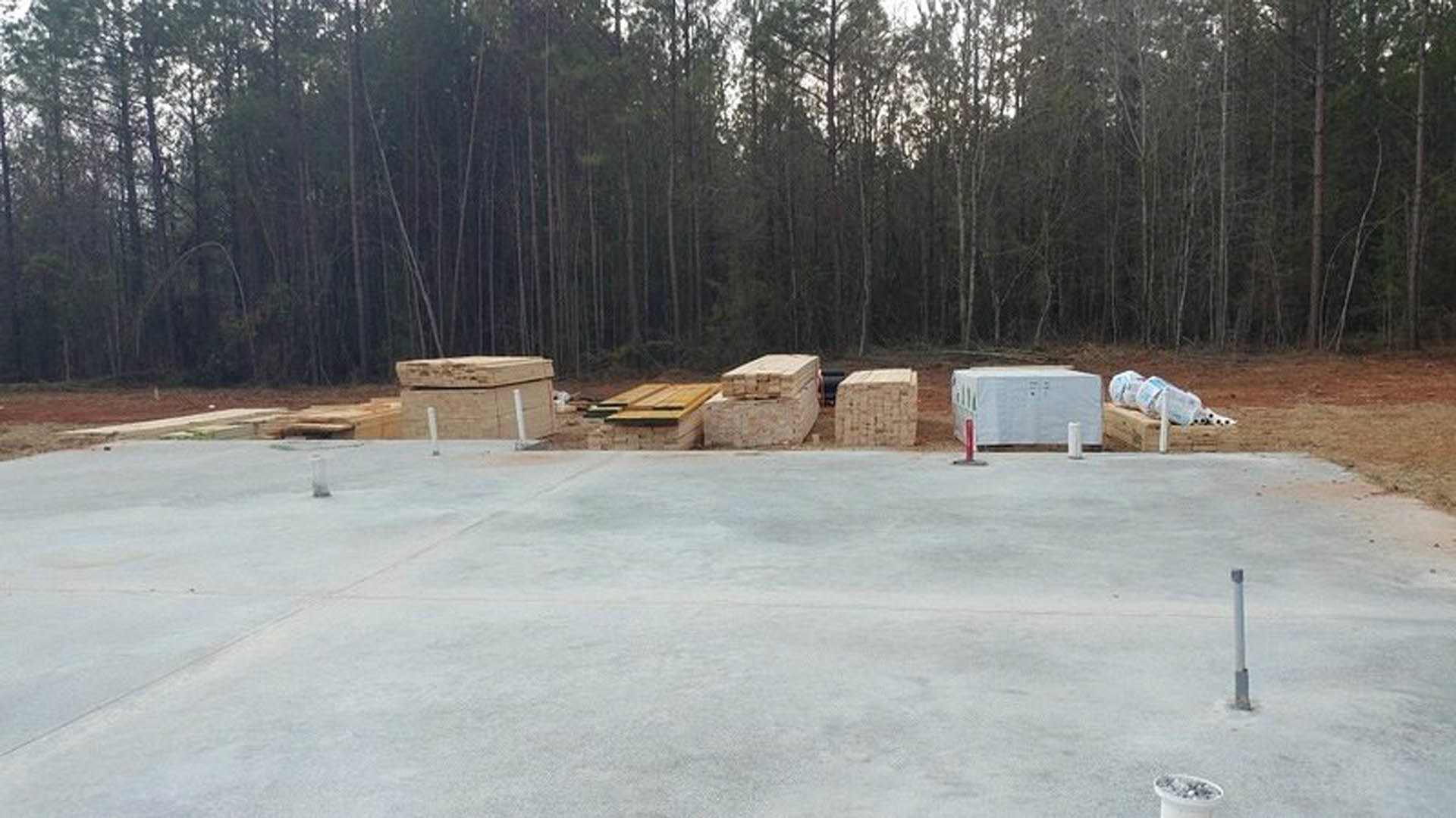 Construction site with stacked wood pallets on bare ground, surrounded by tall trees, adjacent to a brick wall and concrete surface