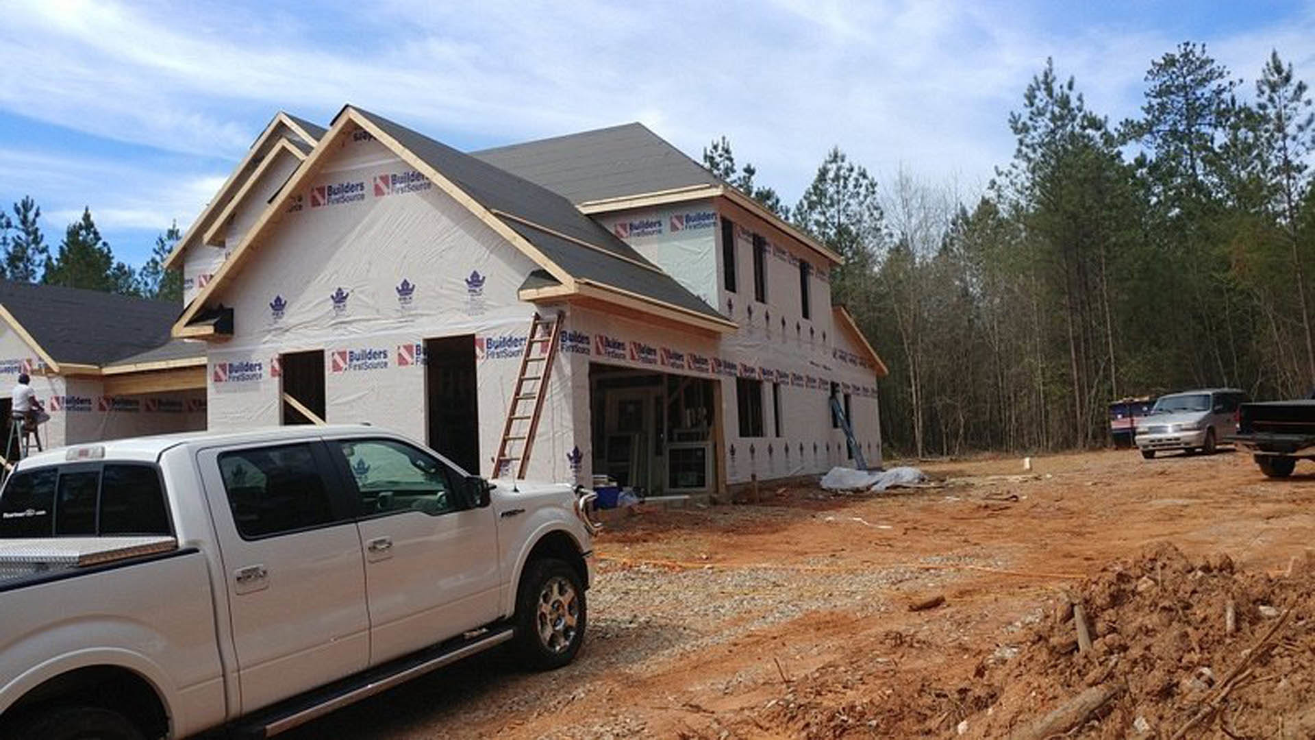 White pickup truck parked on driveway in front of partially constructed house with exposed framing, ladder leaning against exterior, and open truck door visible.