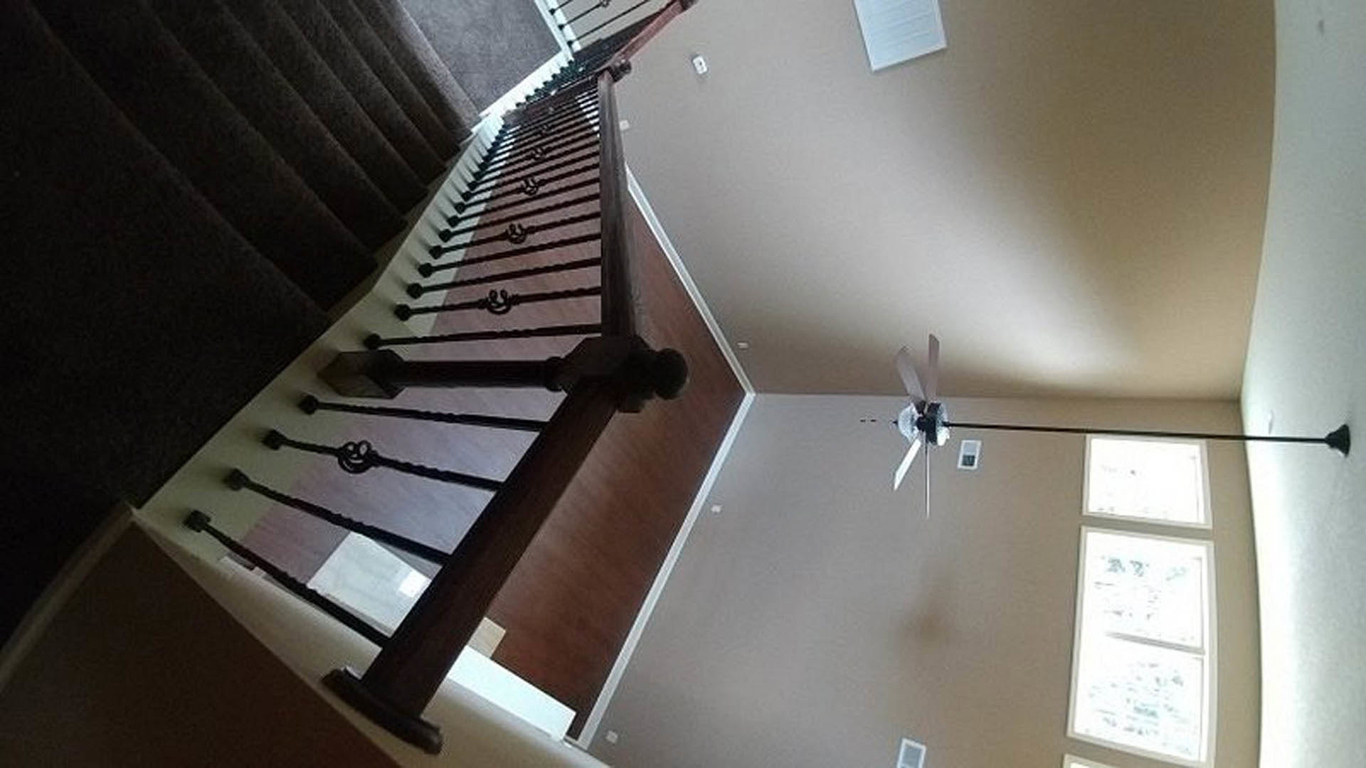 Ceiling fan mounted on white ceiling above staircase with black metal railings, large windows providing daylight, neutral walls, modern interior finishes