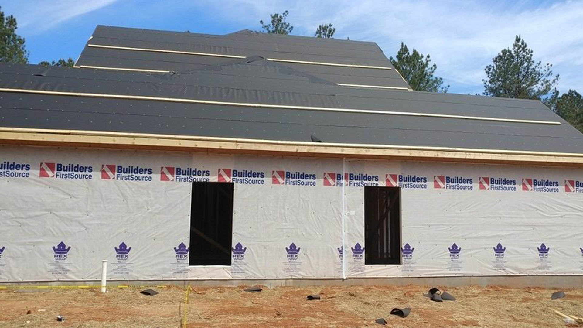 Partially constructed house with black roof, exposed framing, and large window; construction materials and trees visible in background