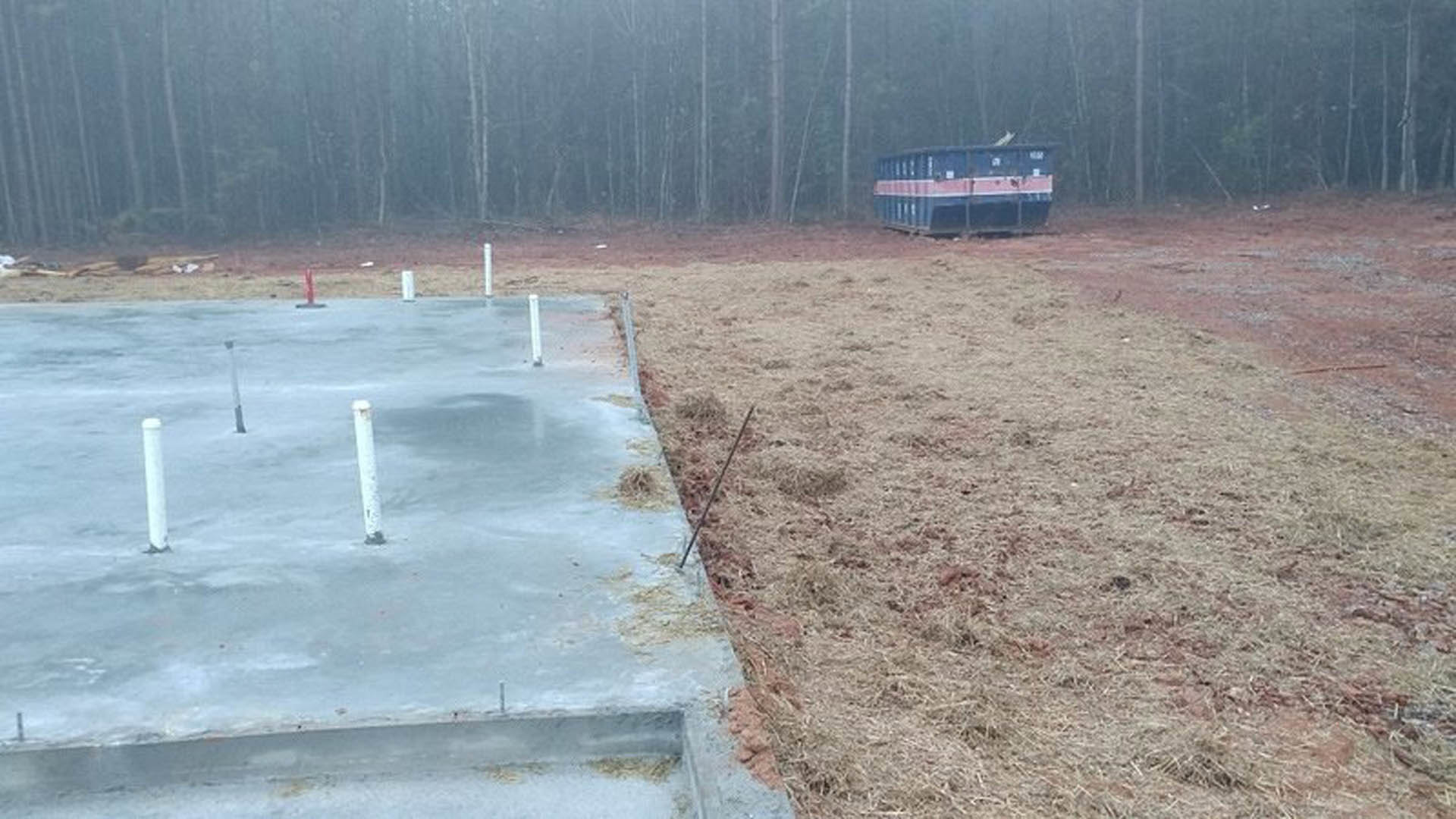 Concrete slab foundation with metal poles, dry grass field, blue and white object in background, foggy trees, person standing in snow