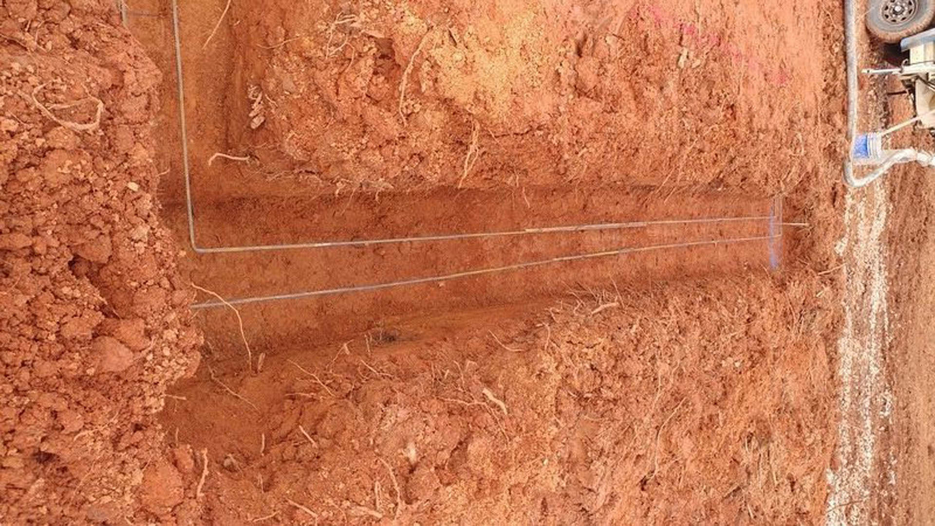 Thin metal wires stretched across a dirt field with scattered small rocks and beige soil, outdoor construction materials visible in the background.