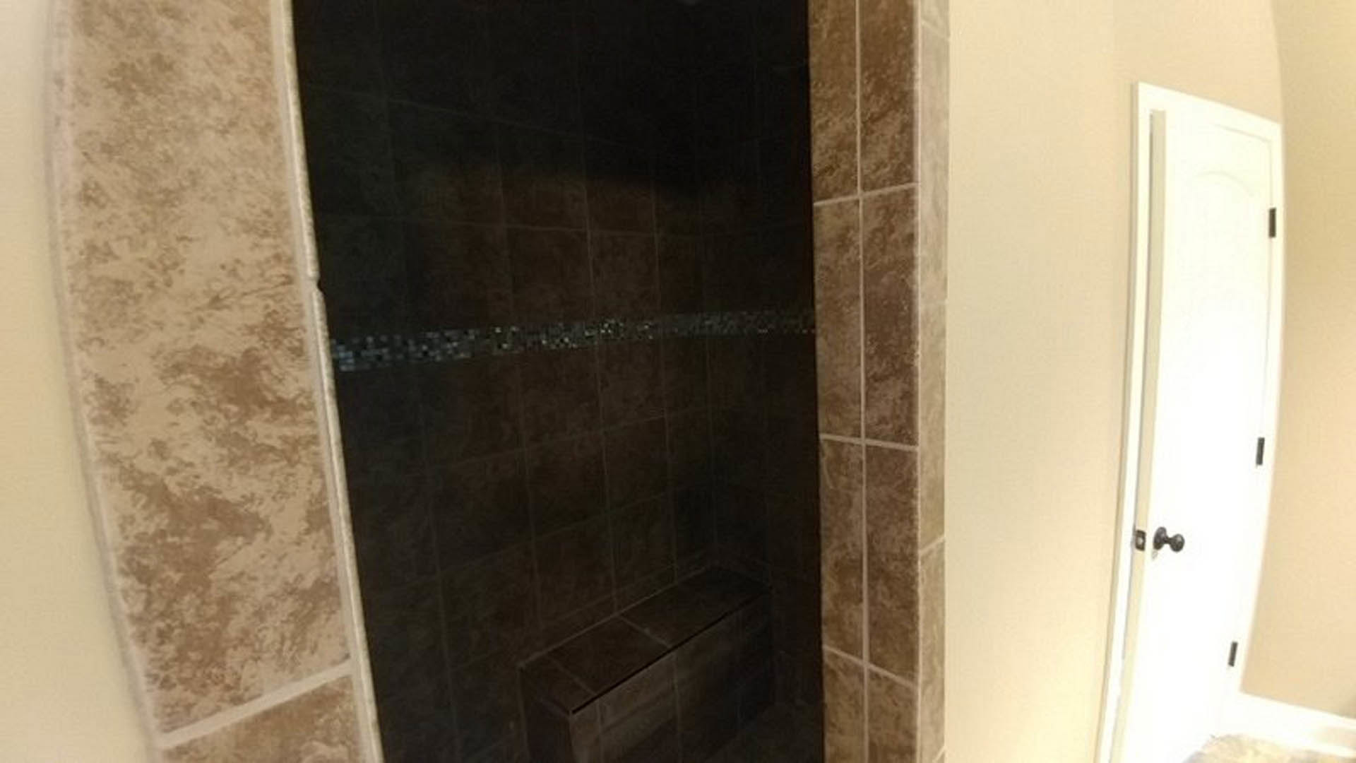 Dark tile shower with contrasting tile border, white door featuring black handle, and plaster walls.