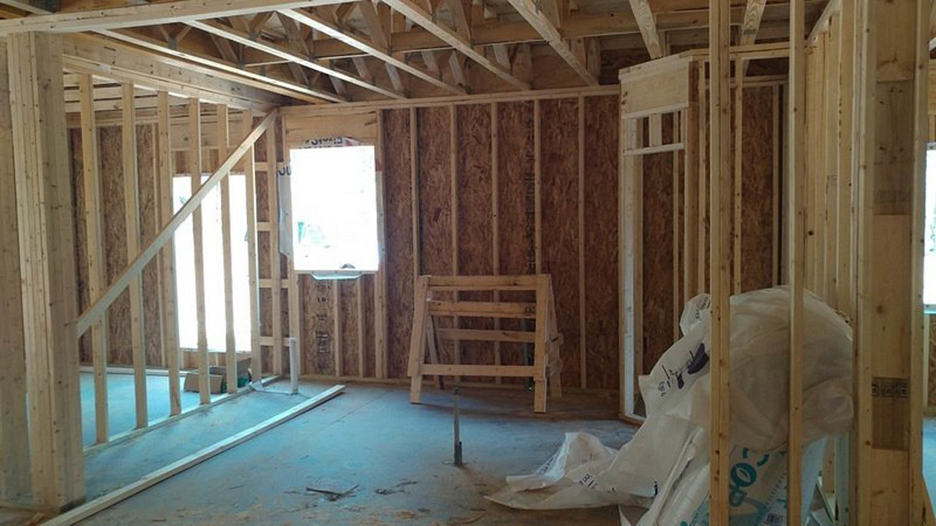 Wood-framed room under construction with exposed beams, insulation, a window letting in natural light, white door, and pile of plastic bags on unfinished floor