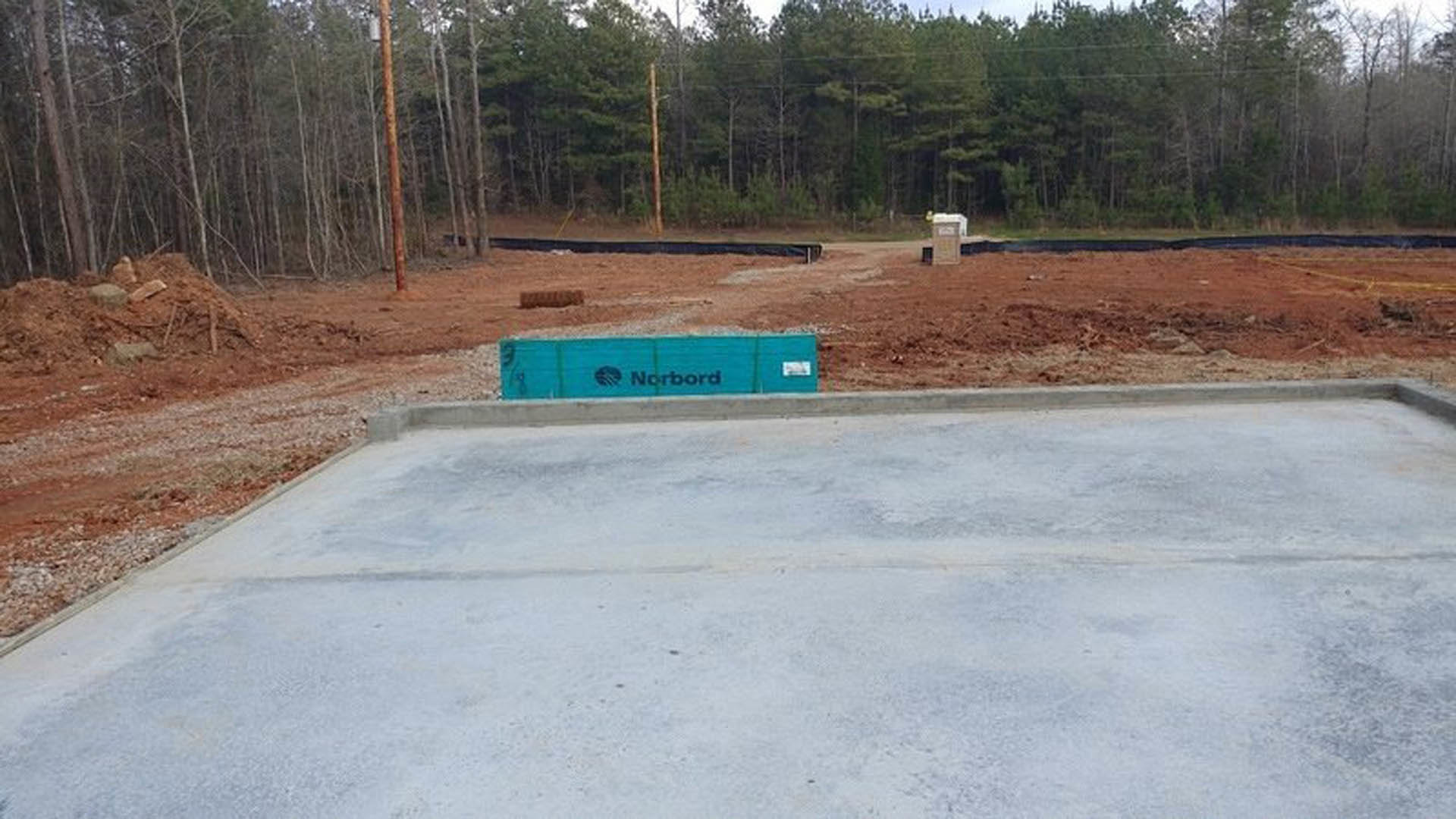 Concrete slab foundation with a blue sign displaying black text, surrounded by dirt and a wooden pole in an outdoor setting with trees and sky in the background.