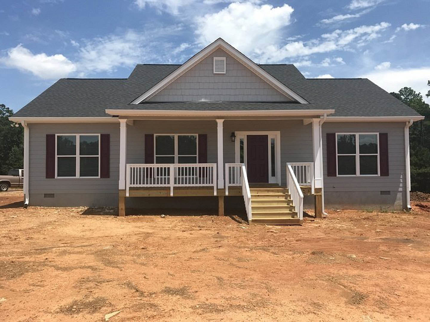Two-story house under construction featuring a covered porch with wooden stairs, black door framed in white, vent on exterior wall, and dirt yard in front.