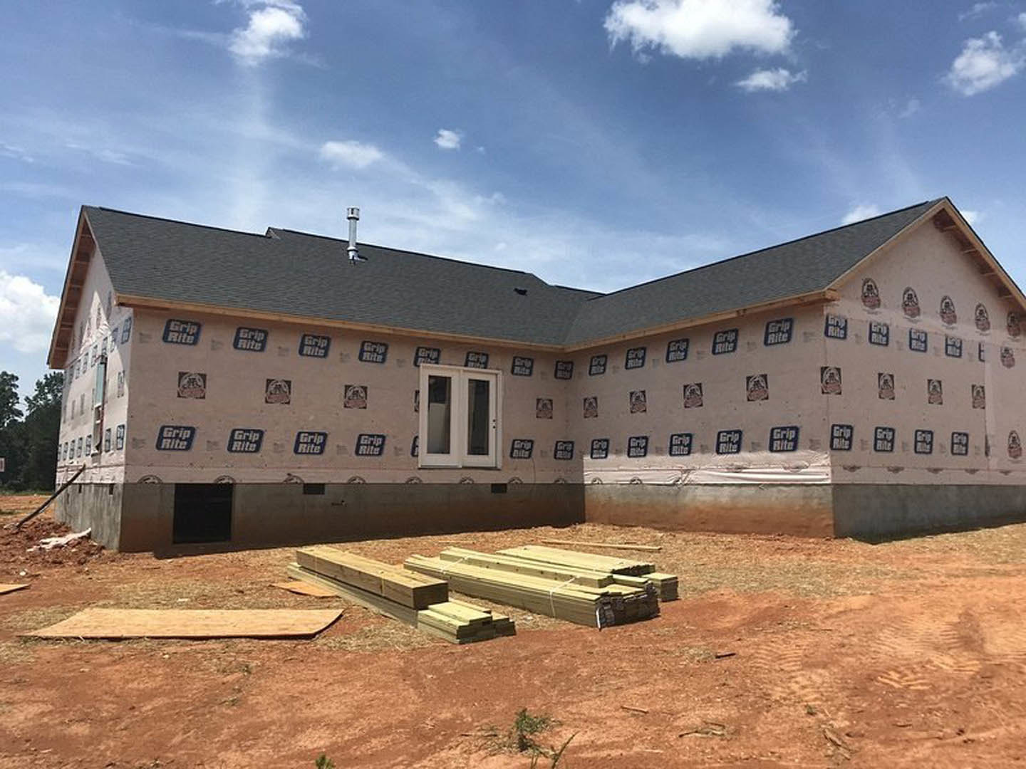 Framed house exterior under construction with exposed wood planks, unfinished walls, and scattered lumber on the ground; partly cloudy sky overhead