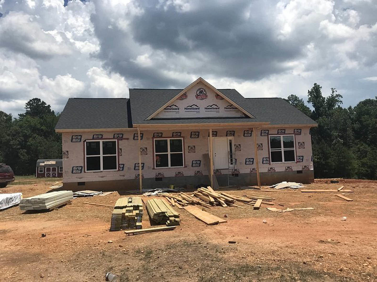 Framed wooden house under construction beneath overcast sky, white beams stacked on a truck, car parked nearby, logo visible on surface