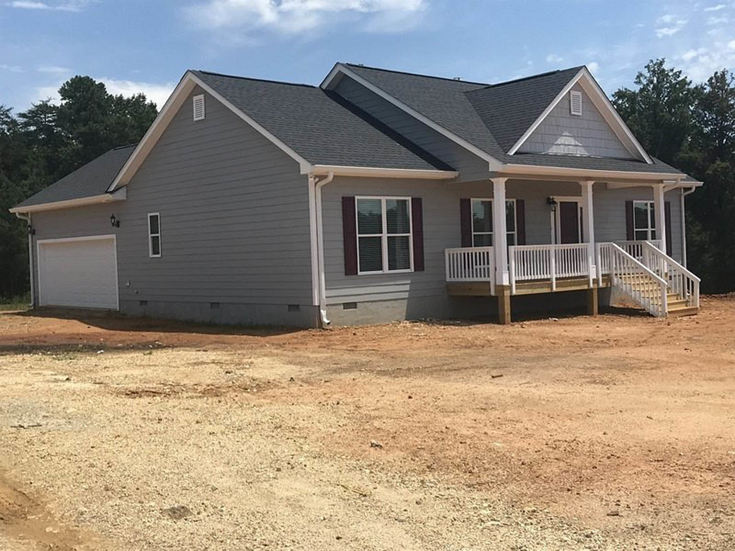 Two-story home with white siding, covered porch featuring white railings, balcony above, white garage door, windows with blinds, dirt driveway, cloudy sky overhead