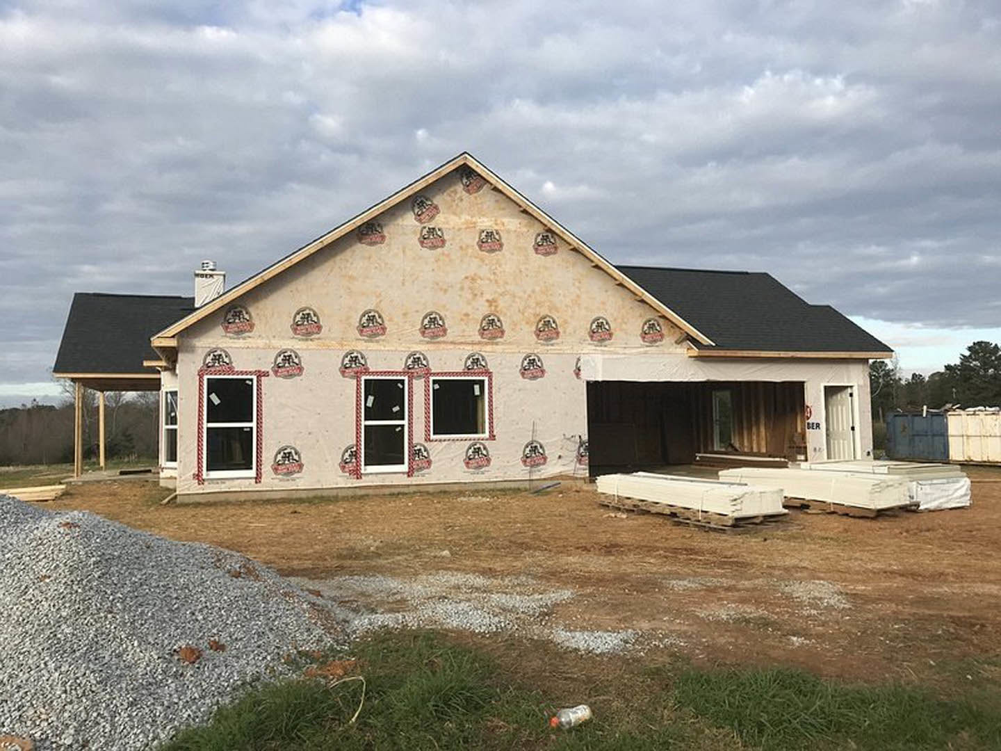 Partially built house with painted exterior wall, white-framed windows, sticker on glass, pile of pallets and gravel on ground, grassy area, cloudy sky overhead