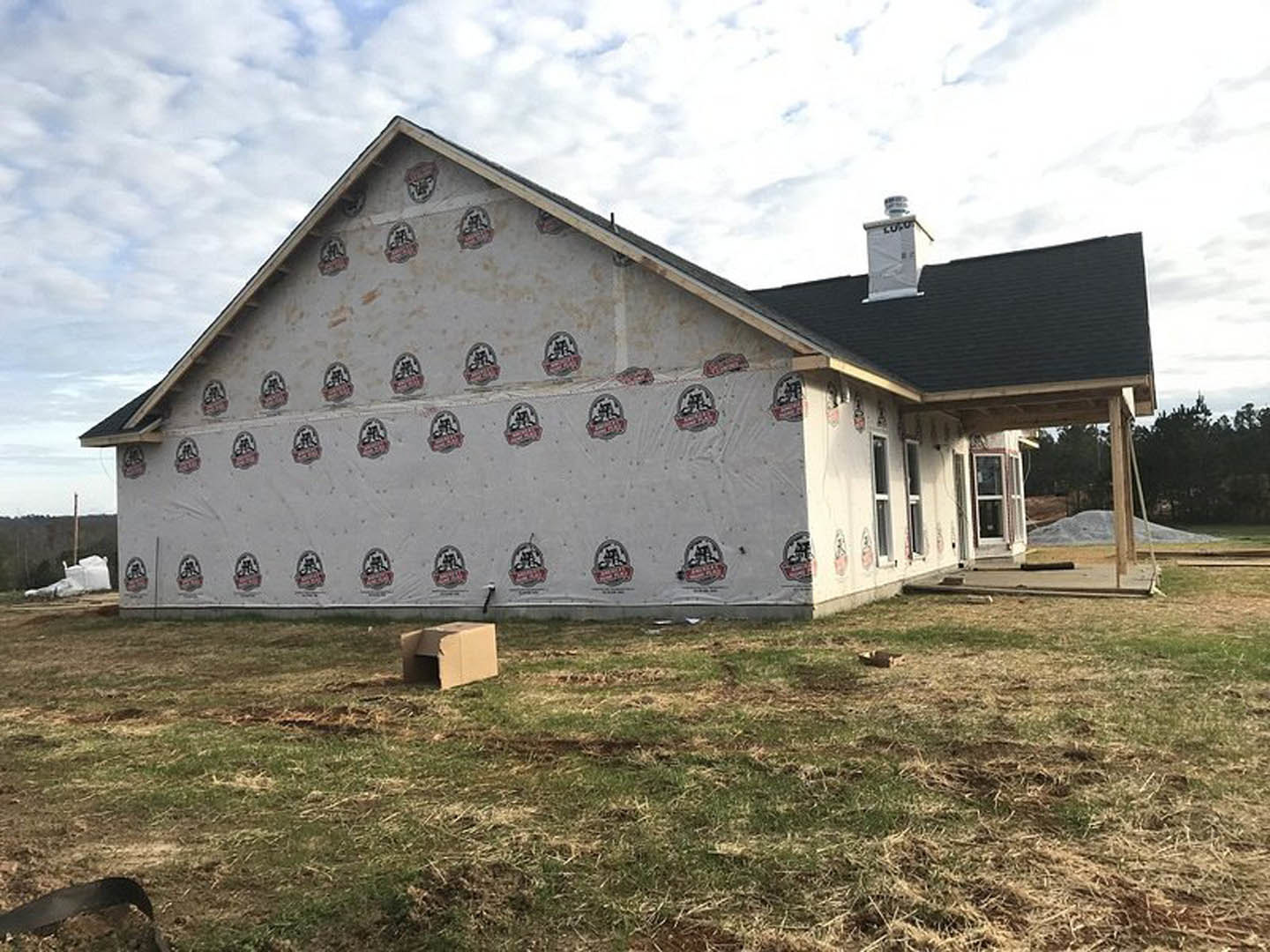 Modern cottage-style home with a covered roof, white exterior walls, black trim, manicured lawn, and surrounding trees under a cloudy sky