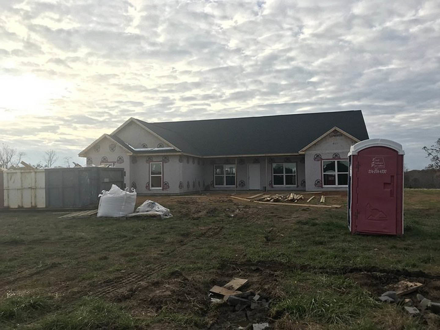 Partially built house with black roof, white-framed window, red waste container, white trash can, and construction materials on grassy lot under cloudy sky