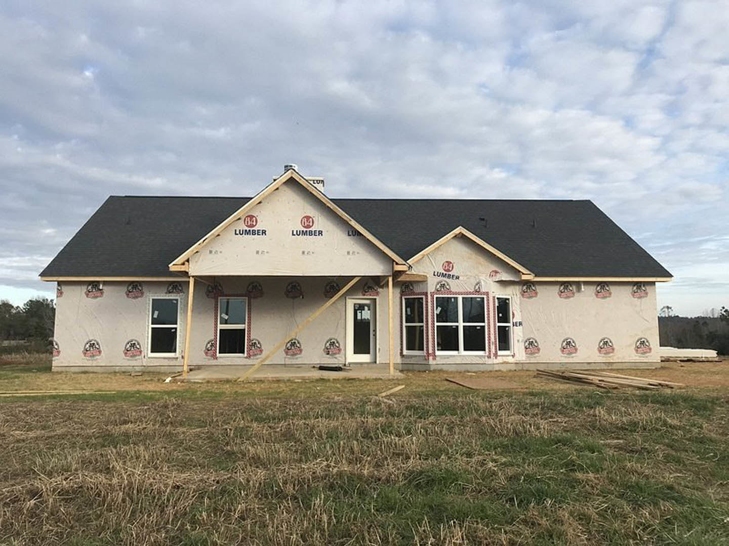 Partially built farmhouse with exposed wood framing, grassy field in foreground, white sign taped to window, cloudy sky overhead