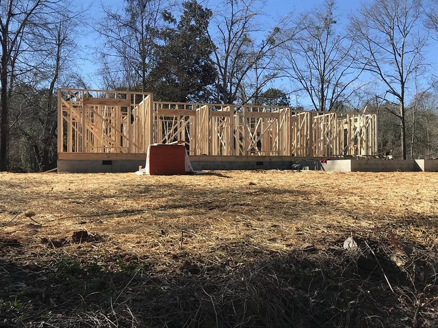 Wood-framed house under construction with exposed beams, fenced perimeter, grassy yard, leafless tree, red toolbox on floor, and red building in background