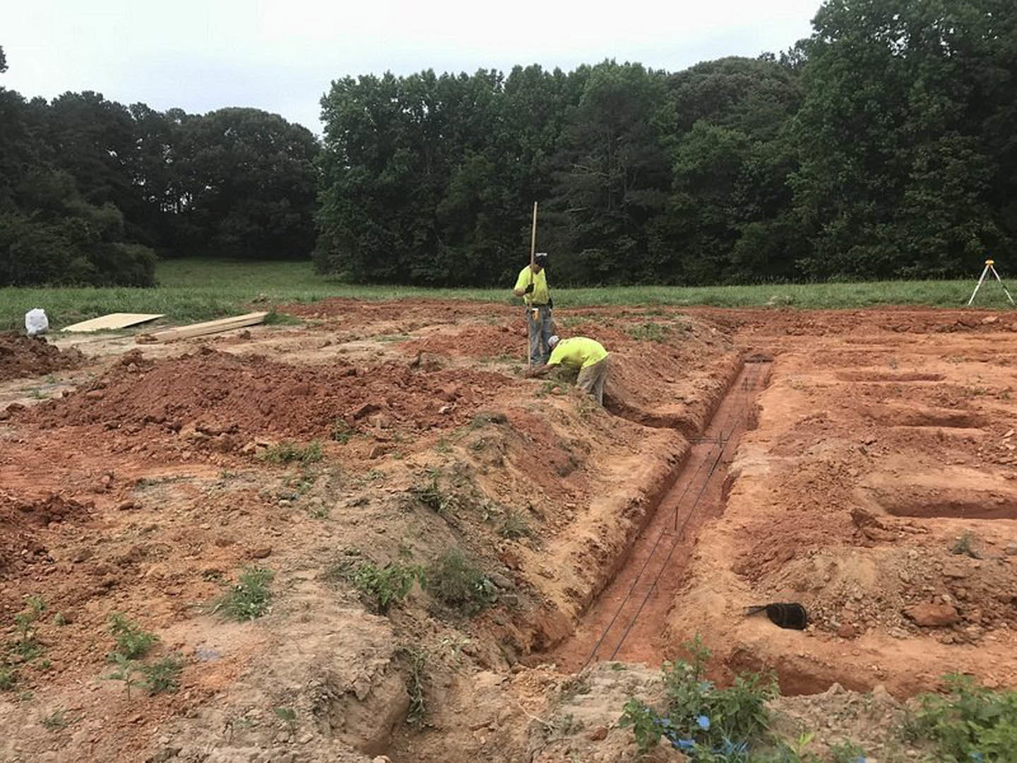 Man standing on dirt field with a measuring pole, surrounded by grass, trees, and open sky