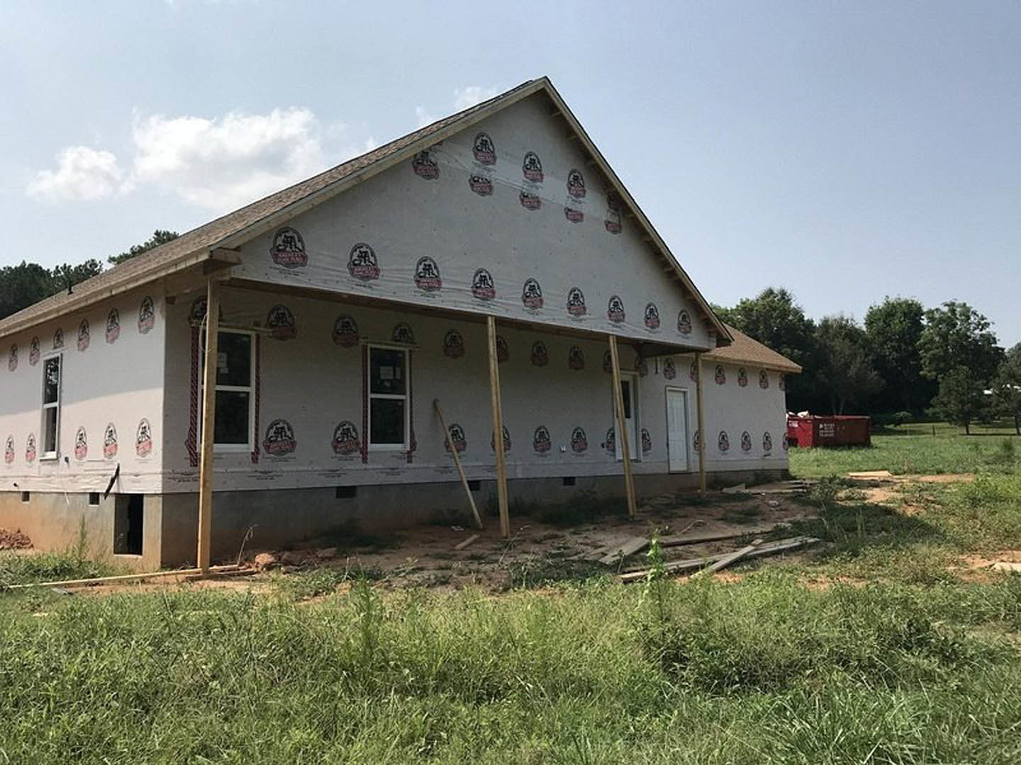 Partially built house with exposed framing, white window frames, construction stickers on exterior, surrounded by green grass and open field under cloudy sky