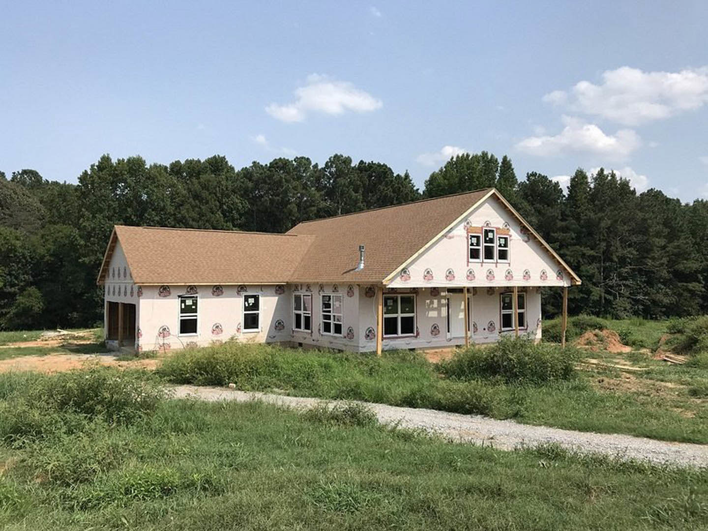 Partially built house with brown roof, white-framed window, exposed pipe, grassy yard, and trees in the background