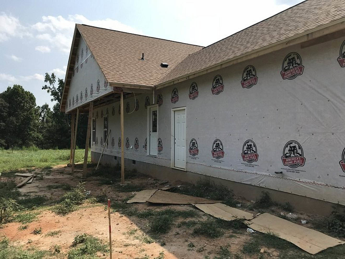 White exterior wall covered in construction wrap with printed logos, black-trimmed white door, grassy lot, mature trees in background, cloudy sky overhead.