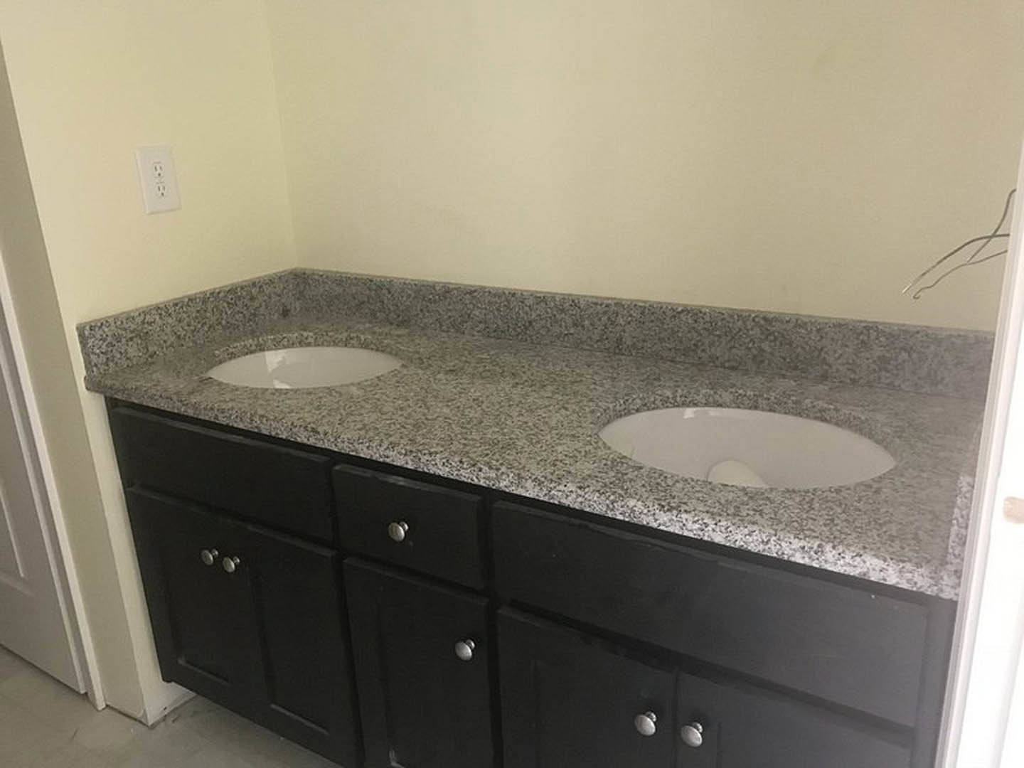 Granite countertop with two white vessel sinks, chrome faucets, tile backsplash, white cabinetry, and electrical outlet visible on the wall