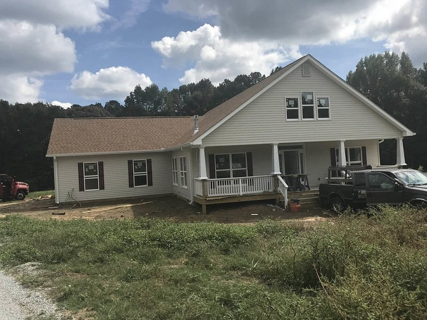 White house with covered porch, white railings, and a black truck parked on the driveway; green lawn and cloudy sky in background.