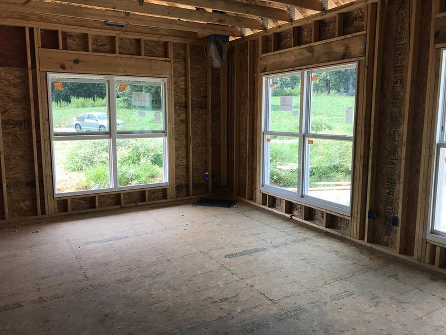Open room with exposed wood ceiling beams, large windows overlooking a green field and parked white car, light-colored walls, and unfinished floor with visible hole.